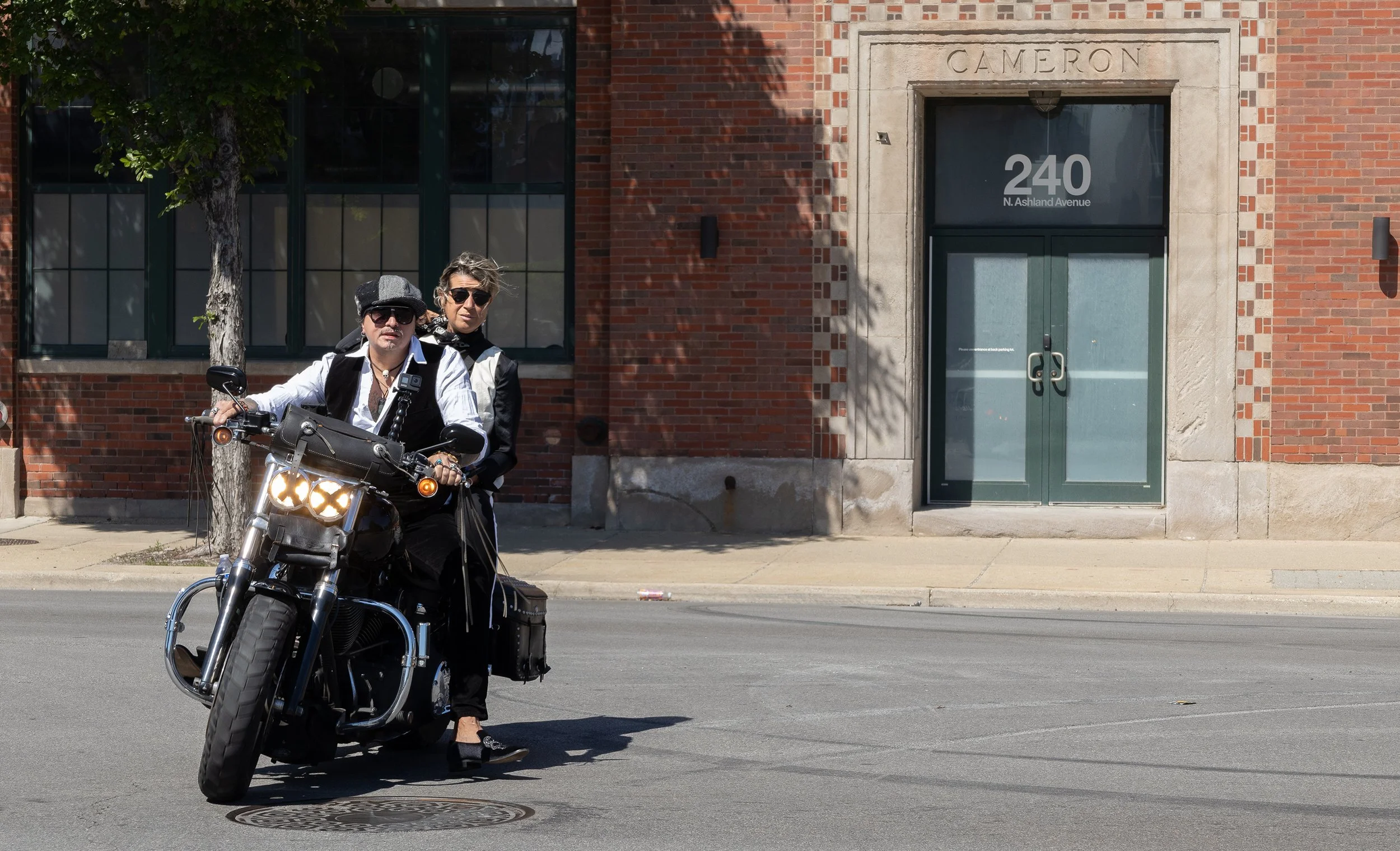 A man and woman, both wearing sunglasses, sitting on a motorcycle on a city street sidewalk, with a brick building and green door behind them.