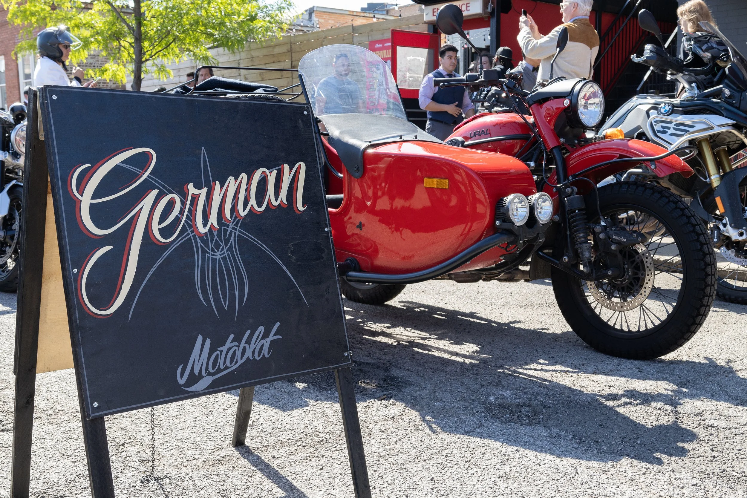 Red motorcycle with a sidecar parked outdoors, with a sign that reads 'German' and mentions a motobalat event. Several people are standing nearby, some taking photos. Trees and a building are visible in the background.