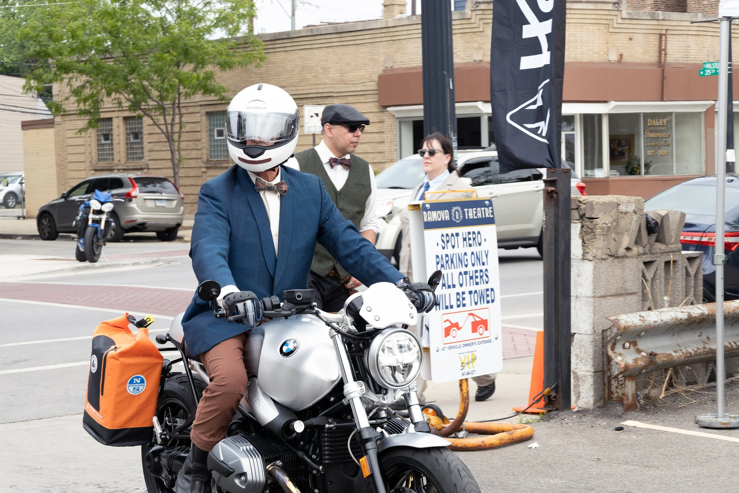 Man wearing a helmet, sunglasses, and a blue blazer riding a BMW motorcycle with orange bags attached on the side, on a city street with a sign indicating parking rules and a black flag with white text