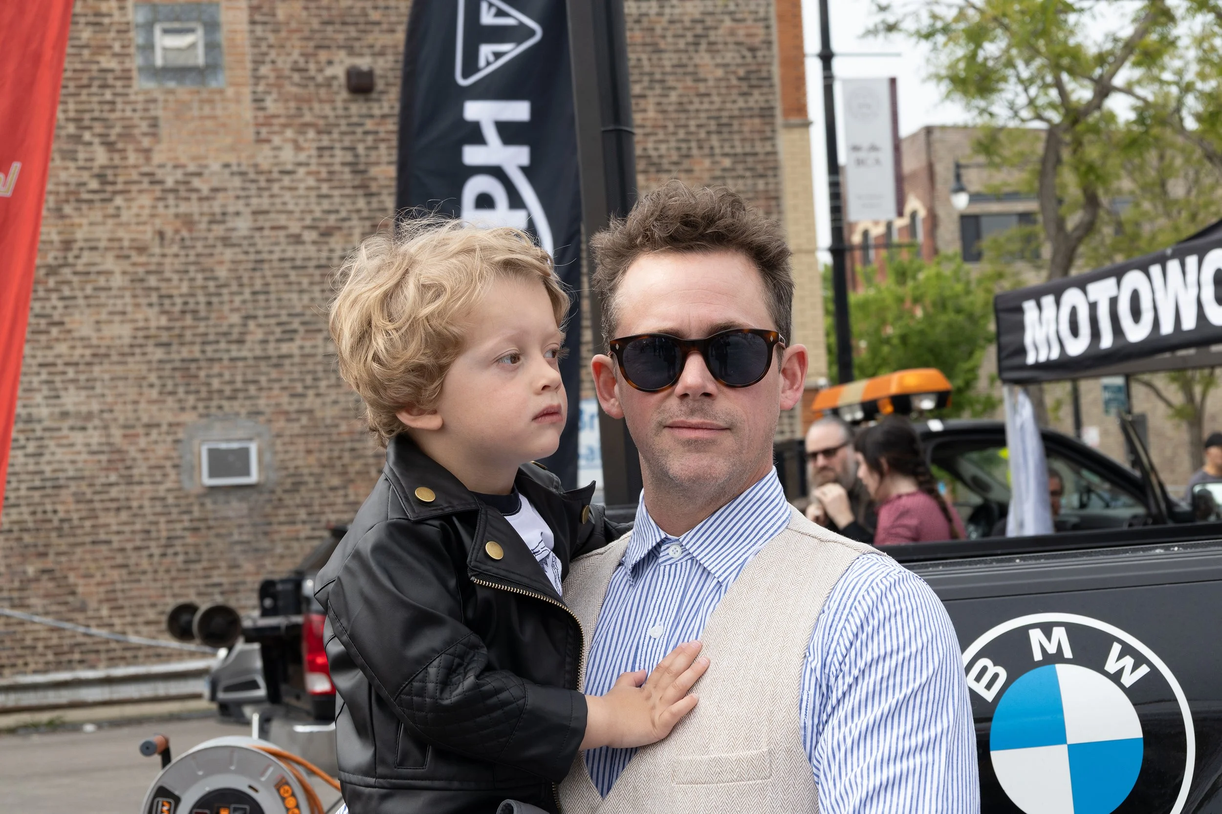 A man with dark sunglasses and a light-colored suit jacket is holding a young boy with blond curly hair and a black leather jacket. They are outdoors in front of a brick building with banners and a BMW logo, in what appears to be a street event or ga