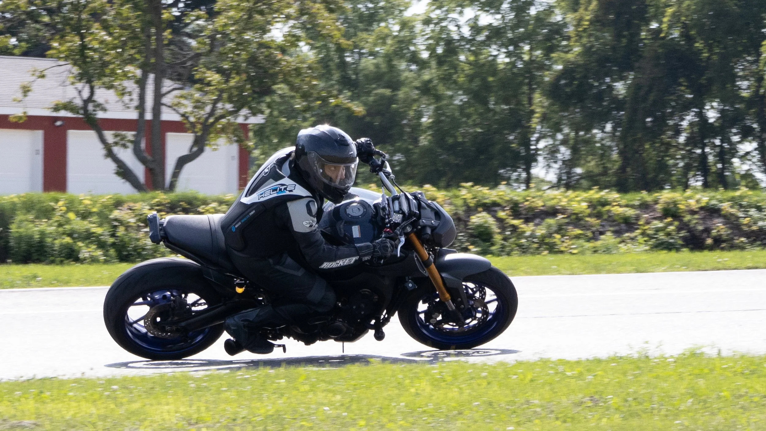A motorcyclist wearing a helmet and protective gear riding a black motorcycle on a suburban street with trees and houses in the background.