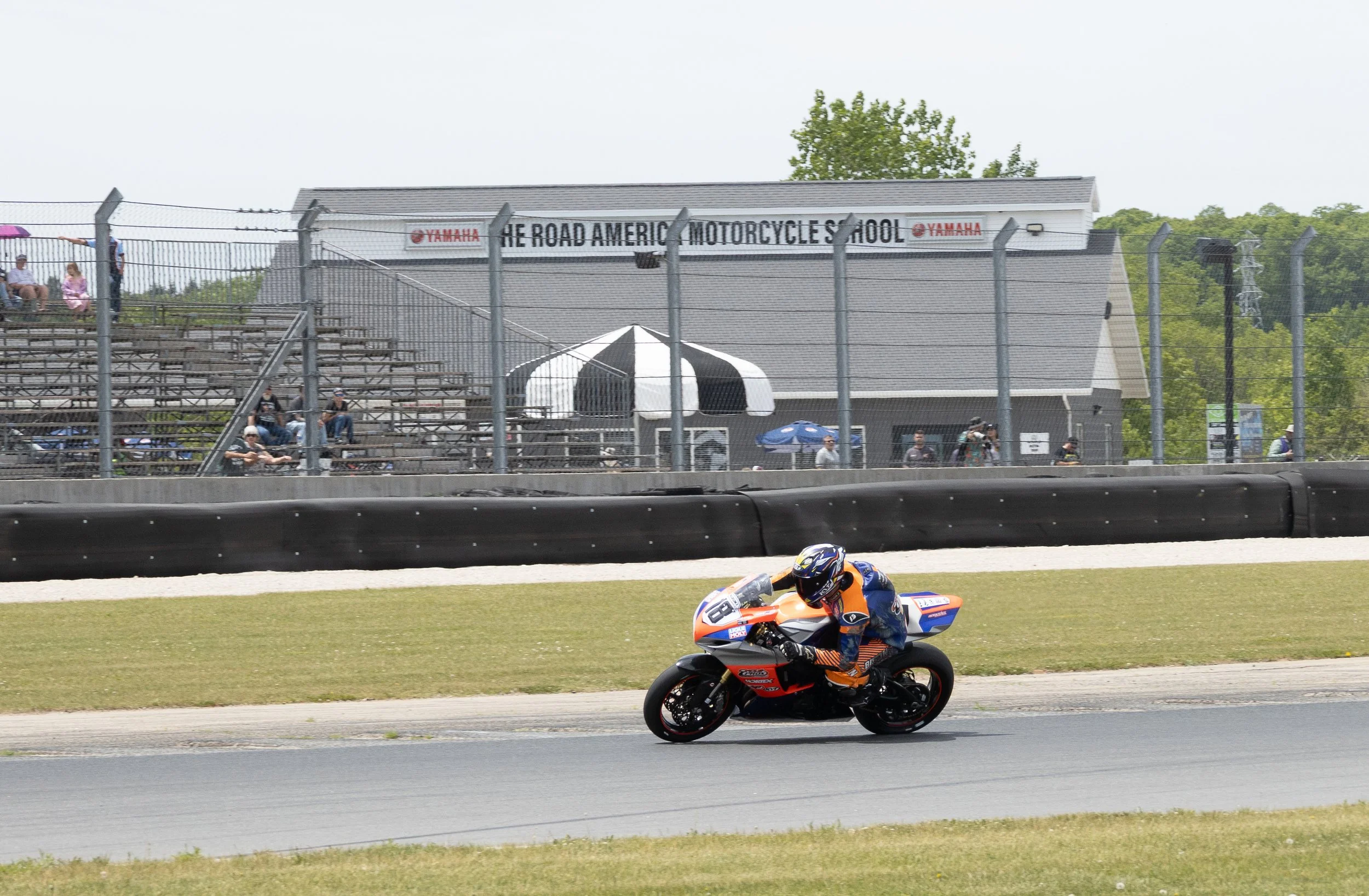 Motorcycle racer in orange and blue gear speeding on a race track with a blurred background of spectators sitting on bleachers and a sign that reads 'The Road America Motorcycle School'.