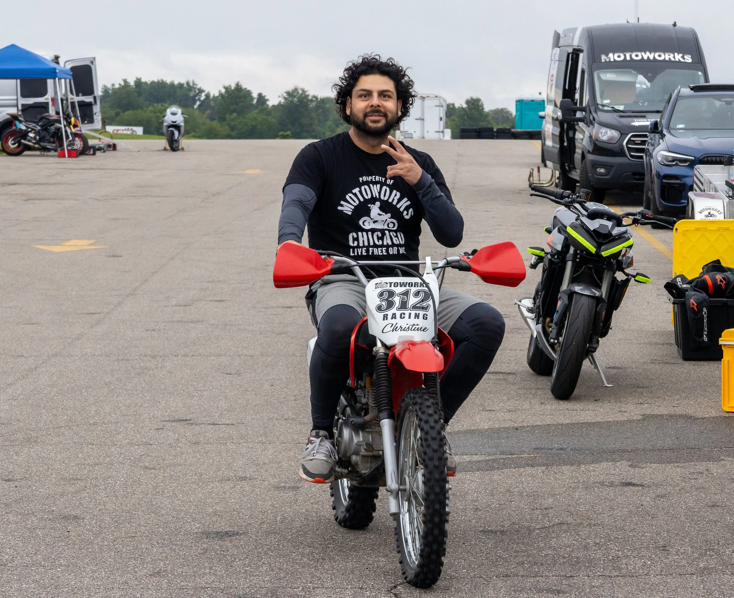A man with curly hair and a beard sitting on a dirt bike in an outdoor parking area, flashing a peace sign, wearing a black t-shirt with white text, surrounded by motorcycles and vehicles under an overcast sky.