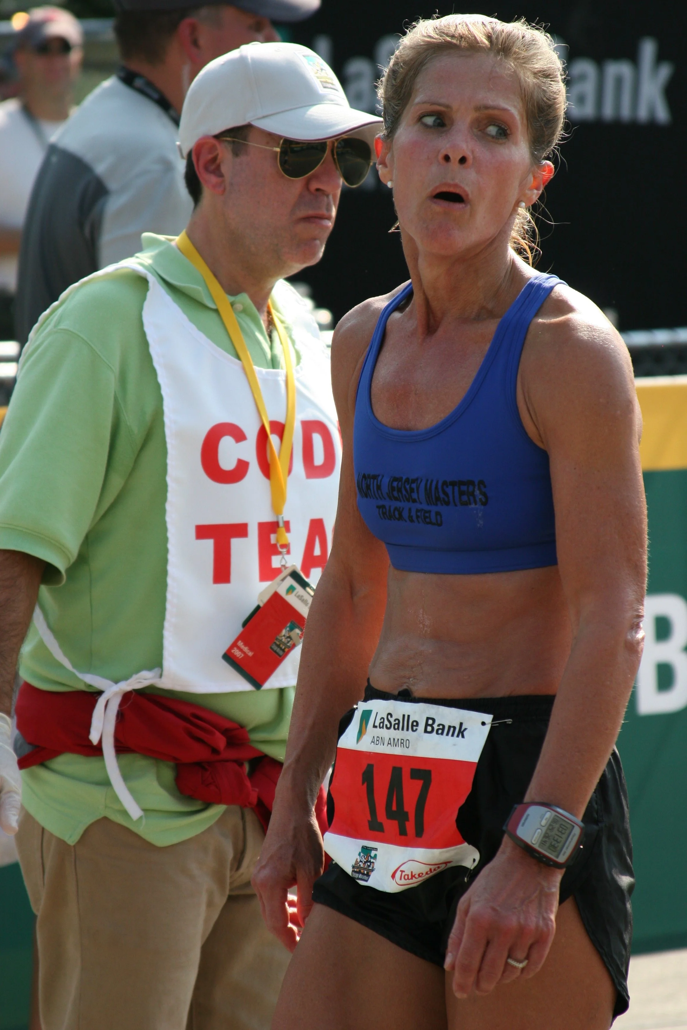 A female marathon runner wearing a blue sports top and black shorts stands with an expression of fatigue or concern, with a race bib numbered 147 and a sports watch on her wrist. An official or volunteer in a green shirt, red shorts, and a white vest