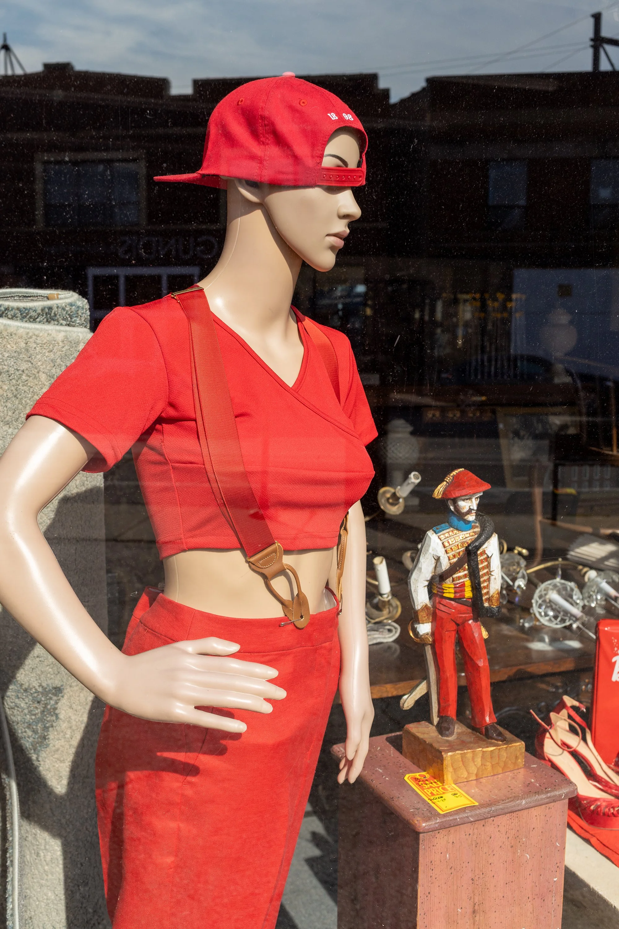 A mannequin dressed in a red crop top and matching red skirt, wearing a red baseball cap backward, displayed in a store window. There are small decorative figurines and a yellow sale sign on a table behind it.