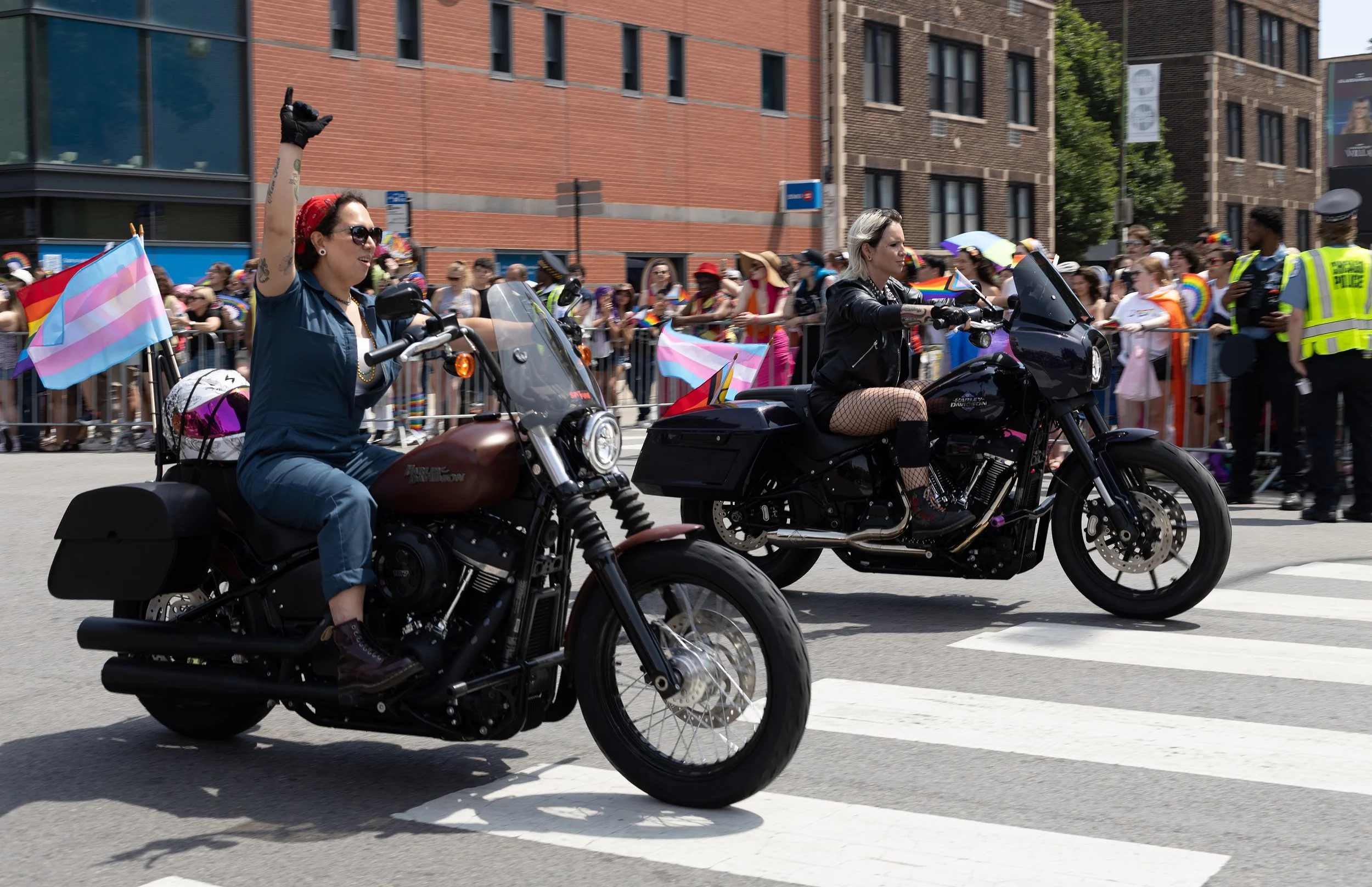 Two women riding motorcycles during a parade, one holding a transgender pride flag. The parade crowd and police officers are visible in the background, with many people holding rainbow or pride flags.
