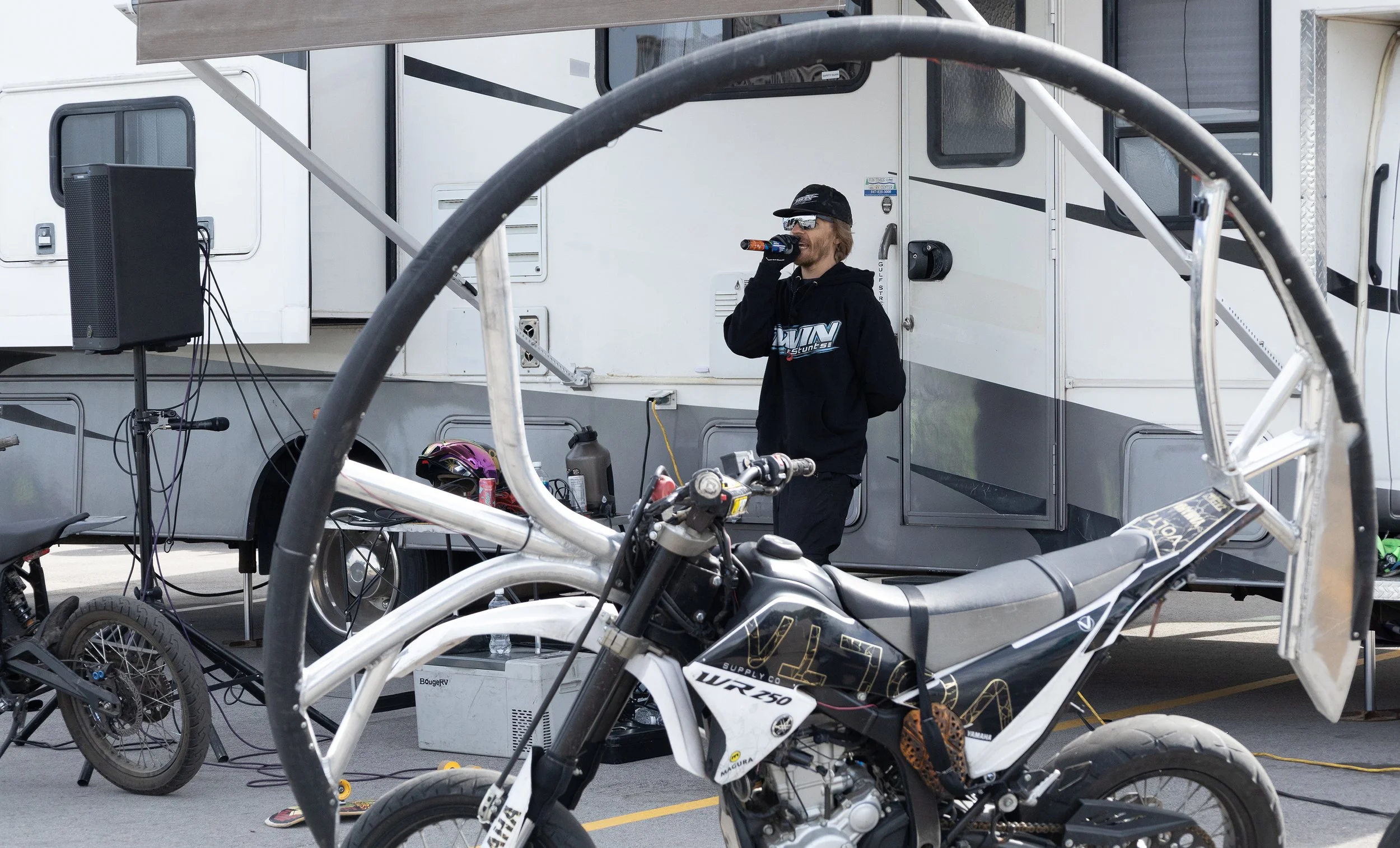 A man wearing a black hoodie, cap, and sunglasses is holding a drink container near his mouth and standing outside a white trailer. There are bicycles and motorcycle parts in the foreground and various equipment around him.