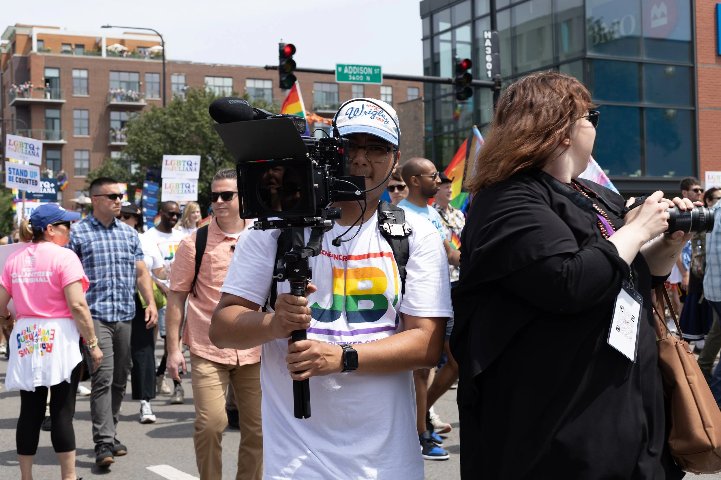 A group of people participating in a pride parade, wearing colorful clothing and holding signs that say "LGBTQ+ JULIANA" and "STAND UP BE COUNTED." Some individuals are carrying rainbow flags. In the foreground, a man is holding a video camera with a