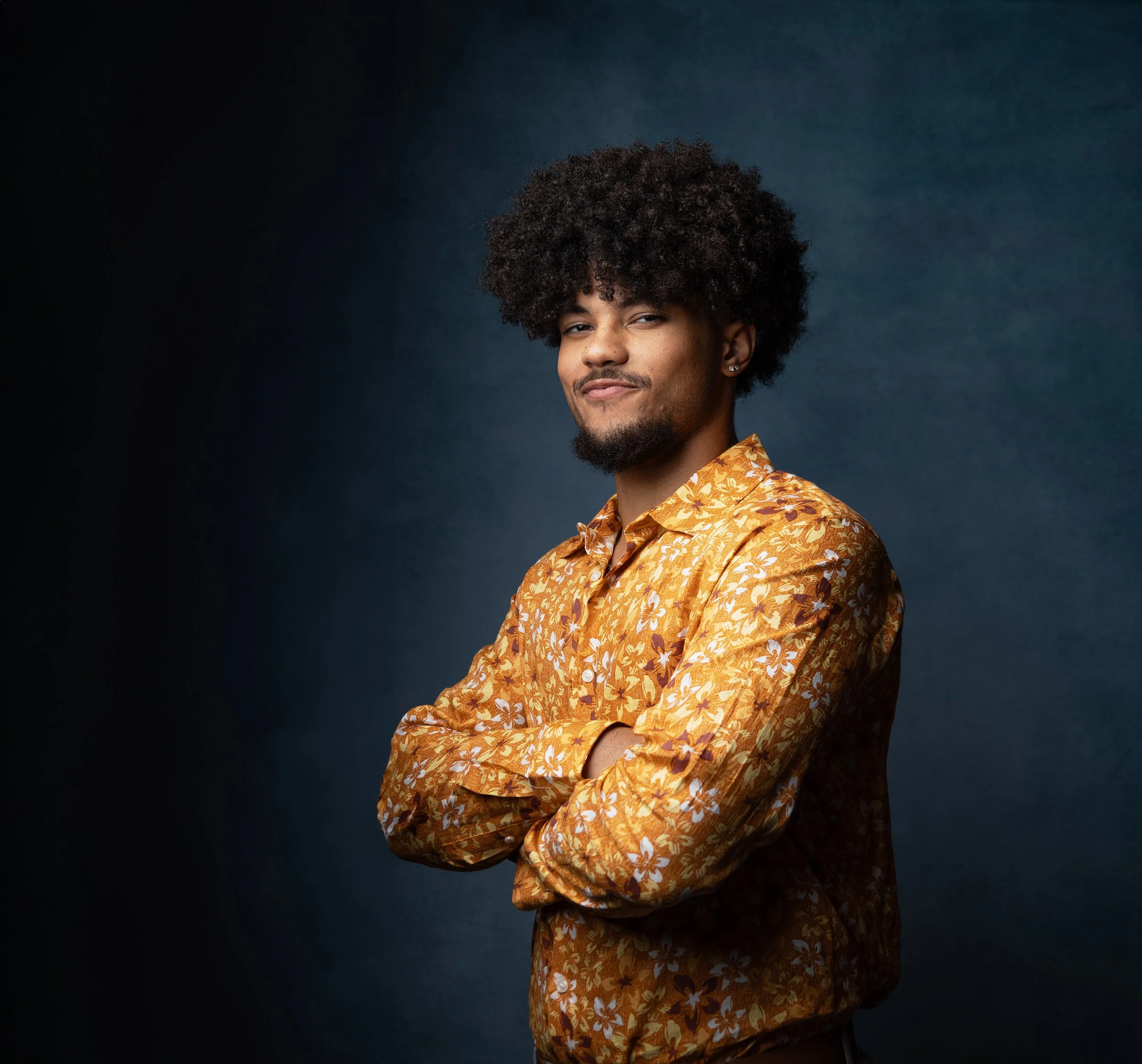 A man with curly dark hair and a mustache, wearing earrings and a yellow floral shirt, standing against a dark blue background.