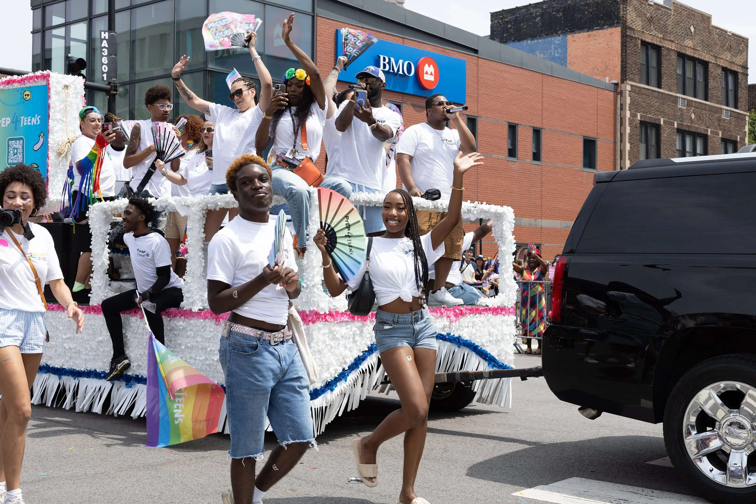 A parade float decorated with white flowers and pink, blue, and yellow accents, carrying a group of diverse people celebrating Pride with rainbow flags and colorful attire. Some are waving, holding signs, or taking photos, as a black vehicle passes b