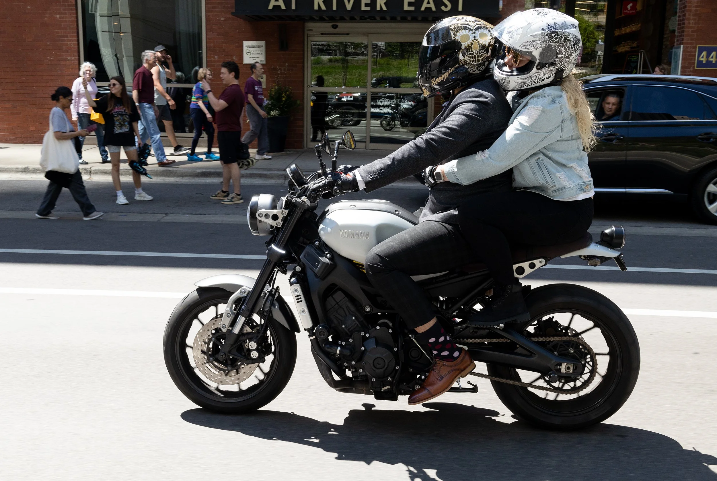 Two women wearing helmets riding a Yamaha motorcycle on a city street. Pedestrians walk past in the background near brick buildings.