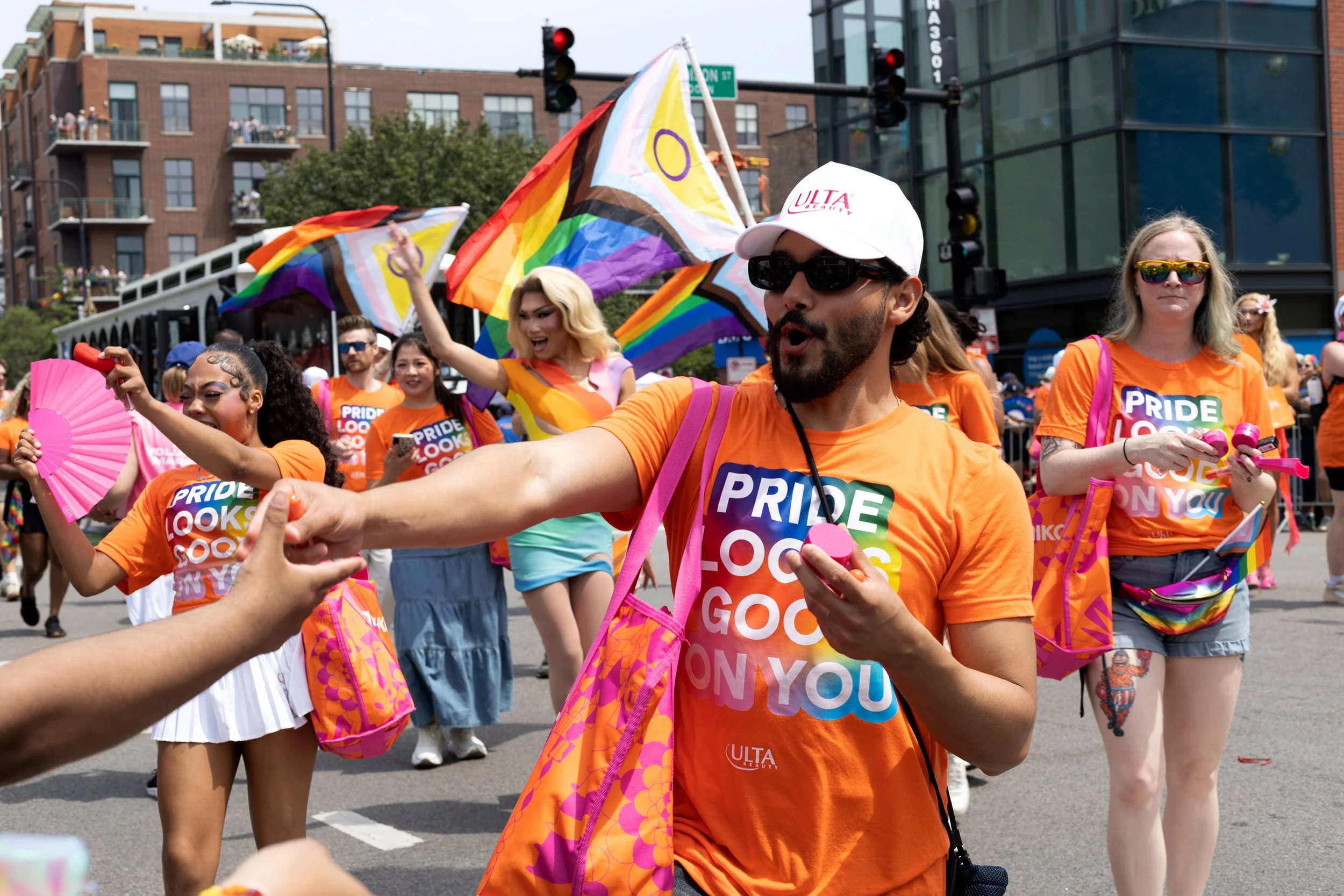 People participating in a Pride parade, wearing orange shirts that say 'PRIDE LOOK GOOD ON YOU', waving rainbow flags and holding pink accessories, with a city street and buildings in the background.