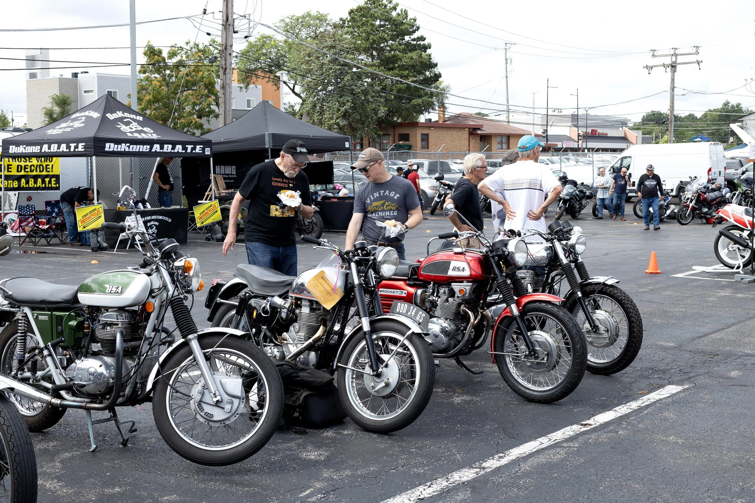 People gathered at a motorcycle event or show in a parking lot with vintage motorcycles in the foreground, tents and parked motorcycles, and a shopping area and street in the background.
