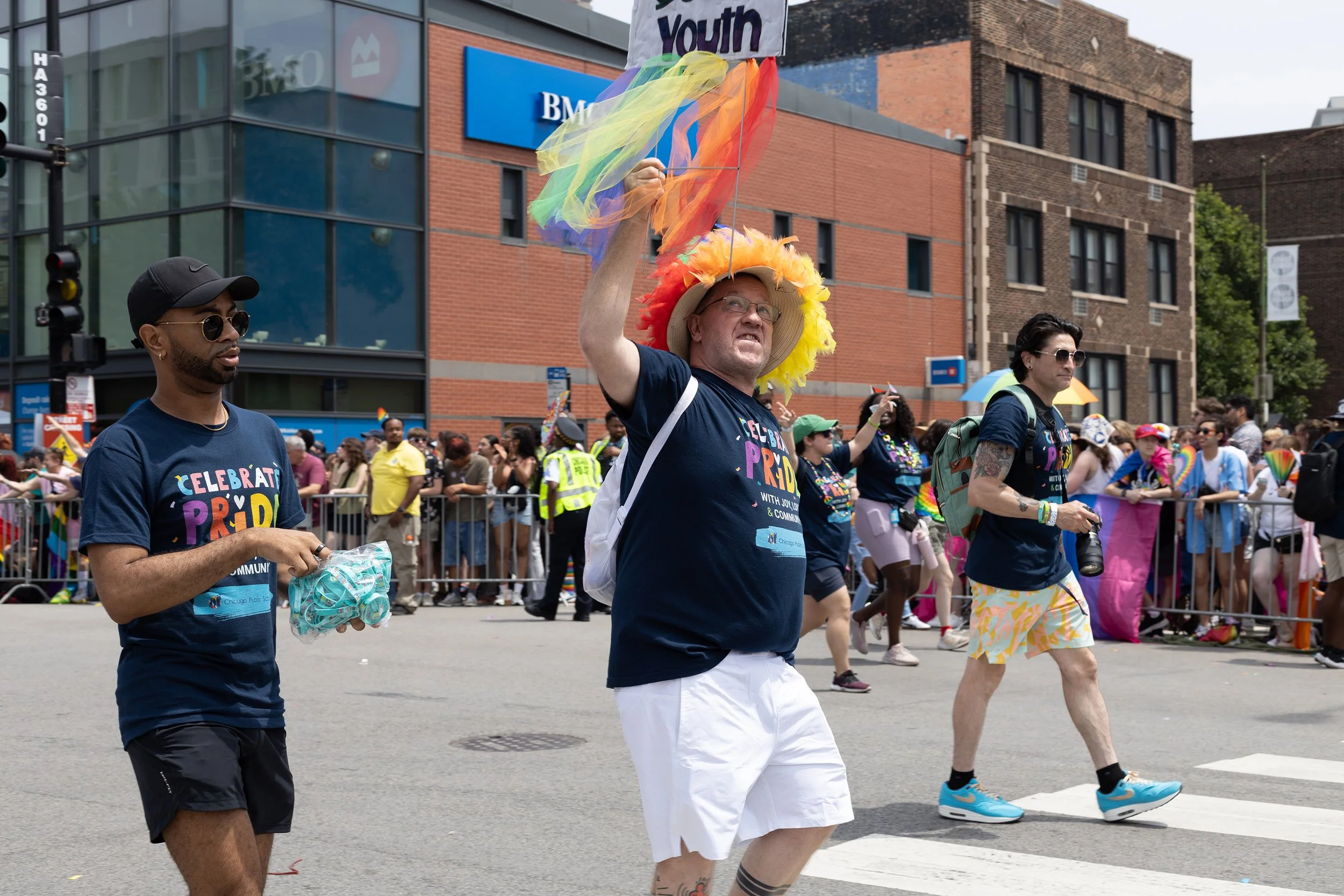 People participating in a pride parade, wearing colorful outfits and accessories. The central figure is a man with glasses and a fancy hat decorated with feathers, holding a rainbow-colored balloon. The background shows a crowded city street with spe