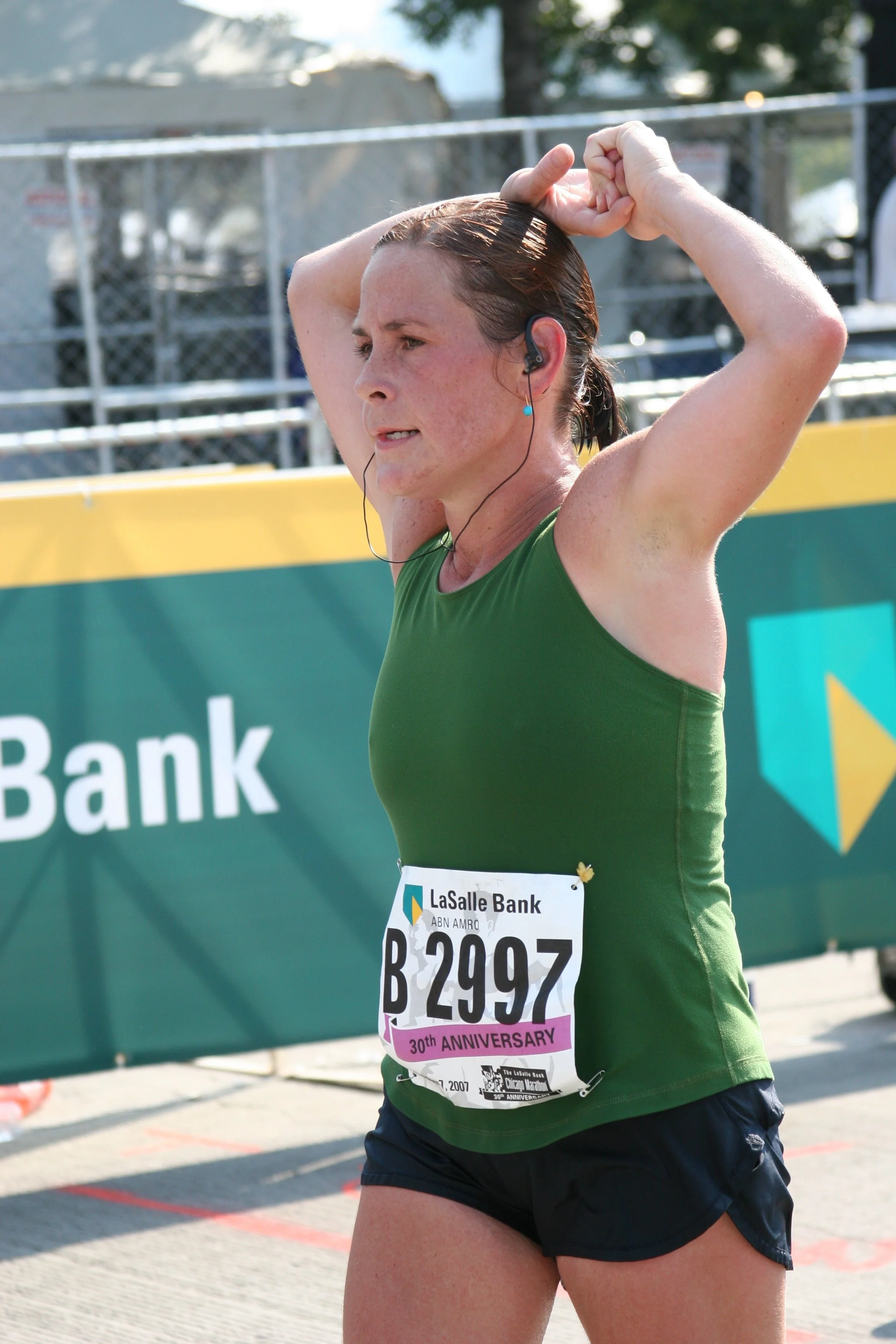 A female marathon runner with her hand on her head, wearing a green tank top, black shorts, and a race bib for LaSalle Bank. She has earbuds in her ears and appears to be at the end of a race.