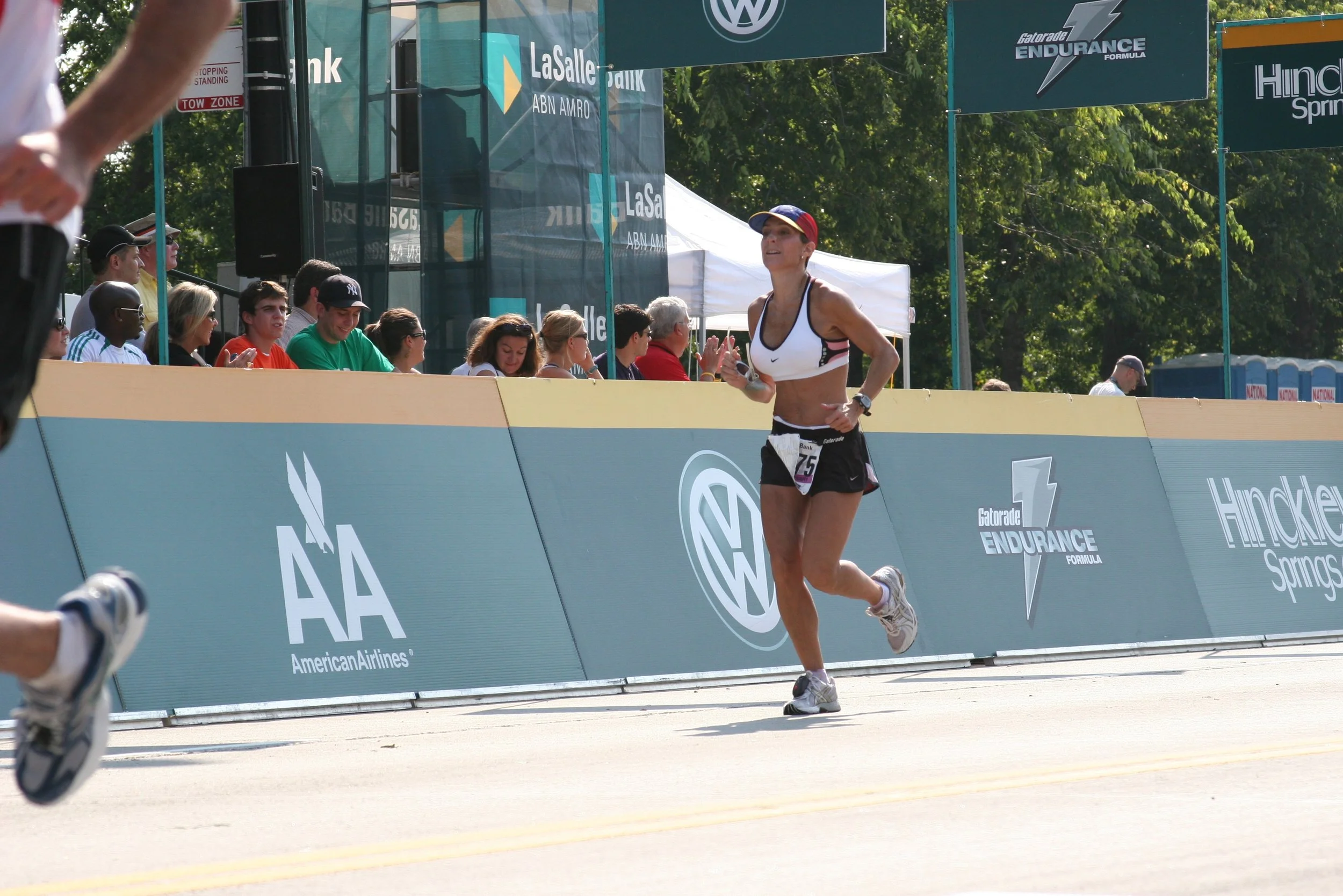 An female runner participating in a race, wearing a white sports bra, black shorts, and a cap, running on a road with spectators seated behind a barrier on the side. There are banners and signs in the background, including logos for VW, LaSalle, Gato