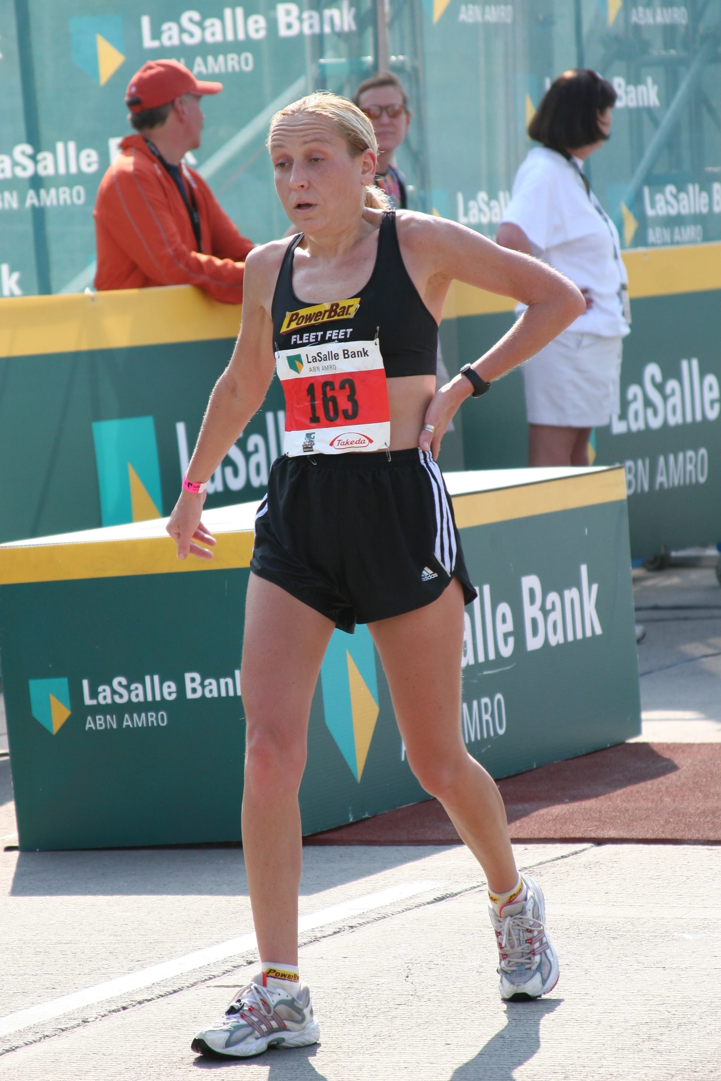 A woman participating in a race, wearing a black tank top, black shorts, running shoes, and a race bib numbered 163. She appears tired or exhausted, with her hand on her side and her eyes half-closed. In the background, there are two women and a man 
