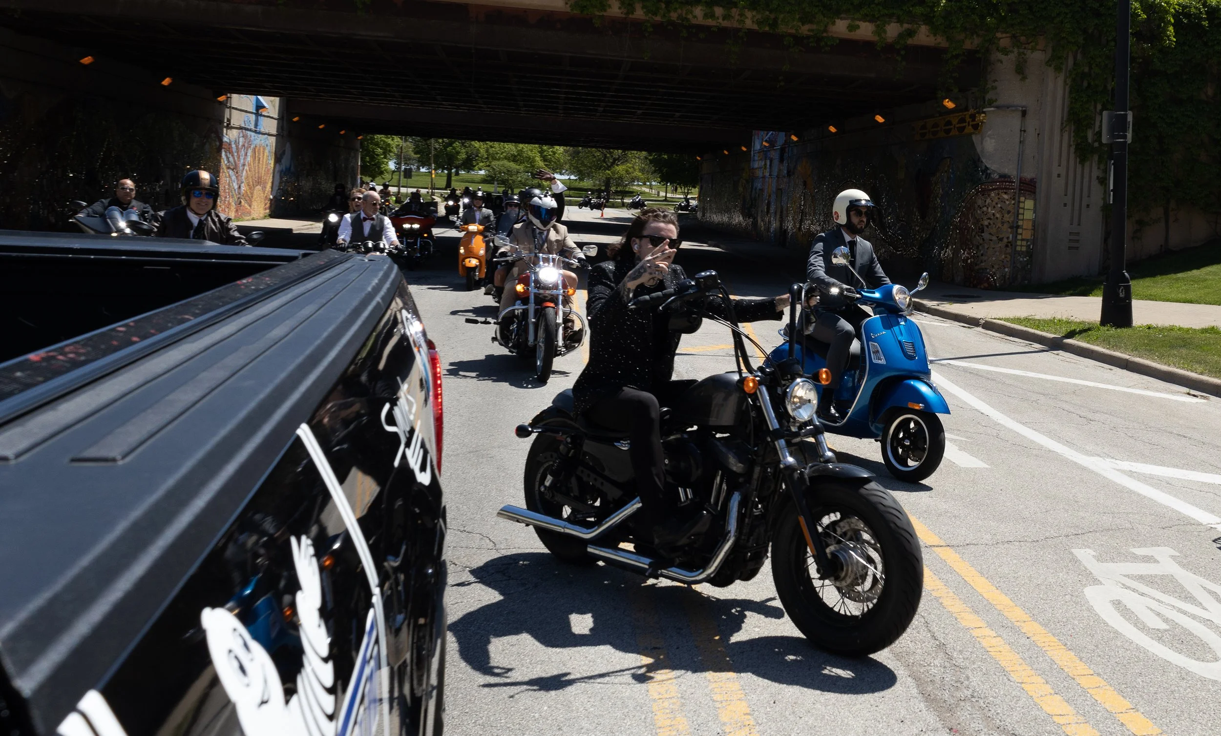 Group of people riding motorcycles and scooters under a bridge on a city street on a sunny day.