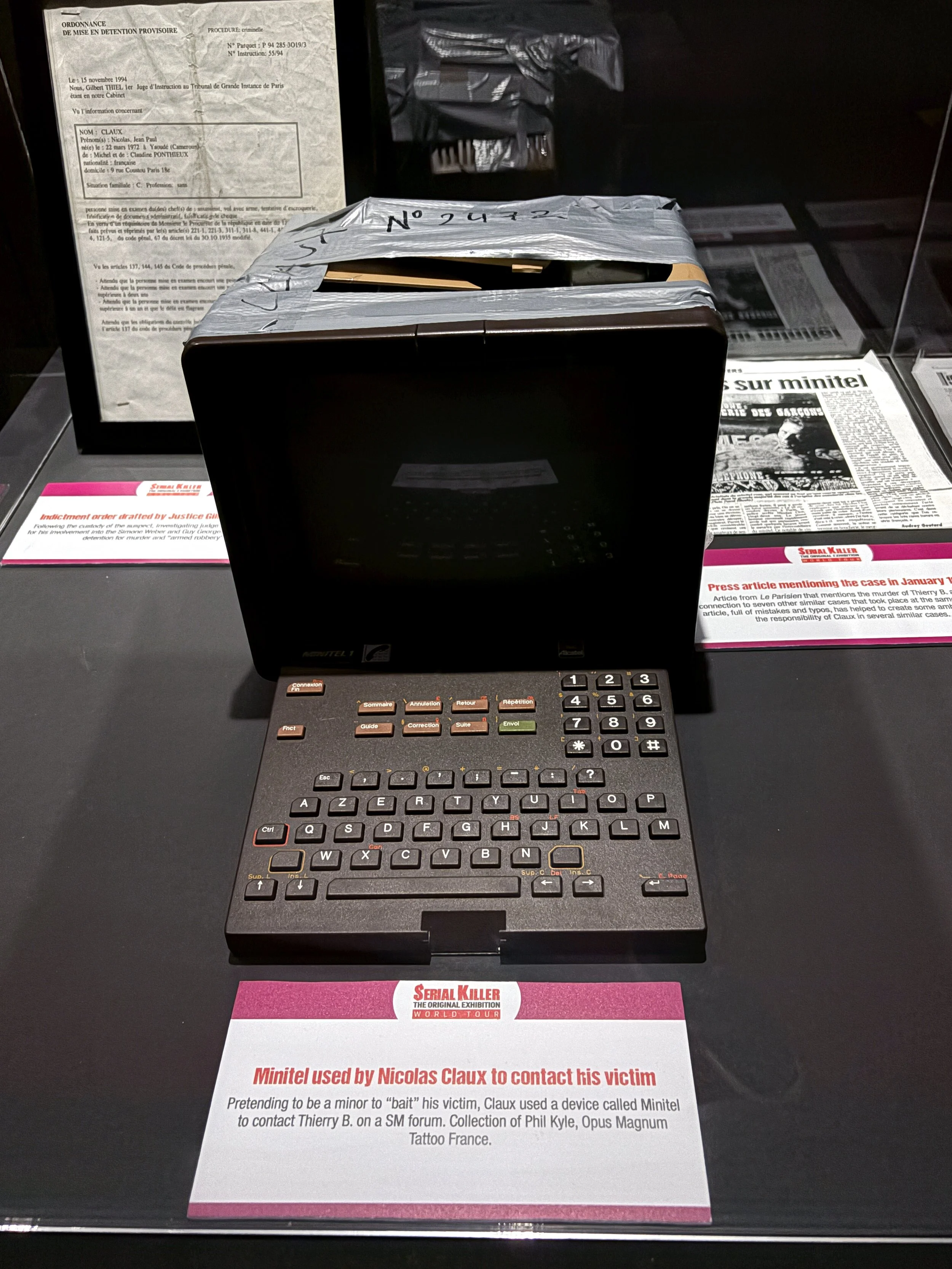 A display case with a keyboard and documents related to a criminal case, including a crumpled piece of tape on a device and informational plaques about serial killer Nicolas Claux and his contact with his victim.