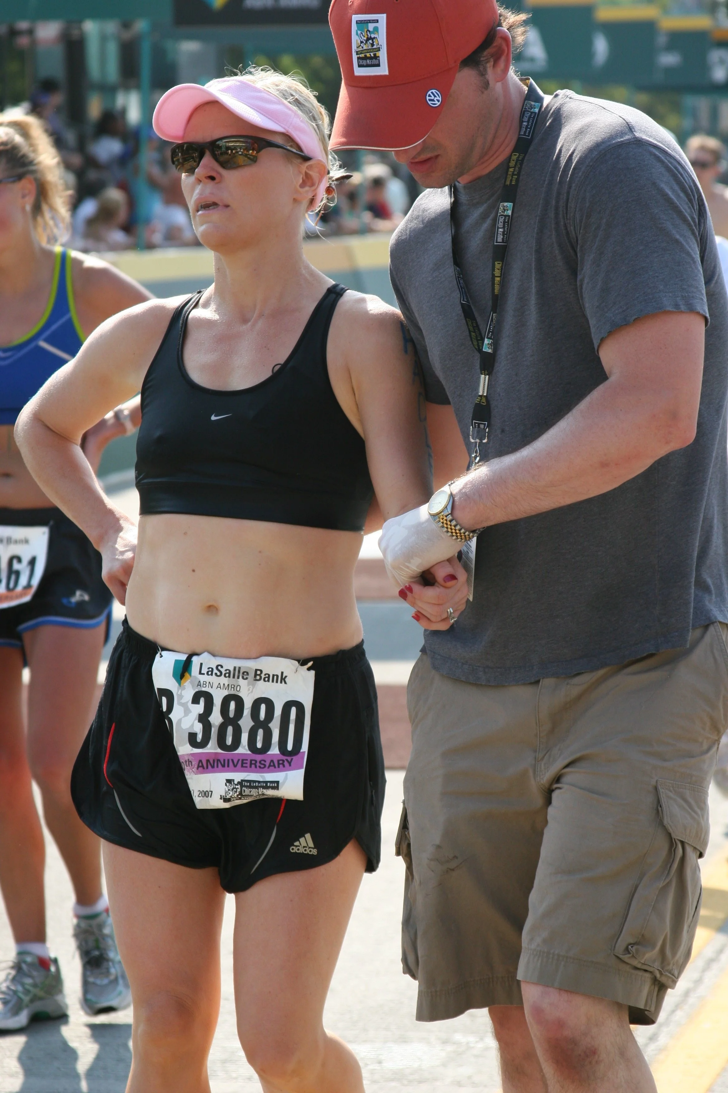 A female marathon runner in black athletic gear with a pink visor and sunglasses is near a male volunteer or coach wearing a gray t-shirt, khaki shorts, and a red hat. The runner has a race bib with the number 33880 and an anniversary logo on it. The