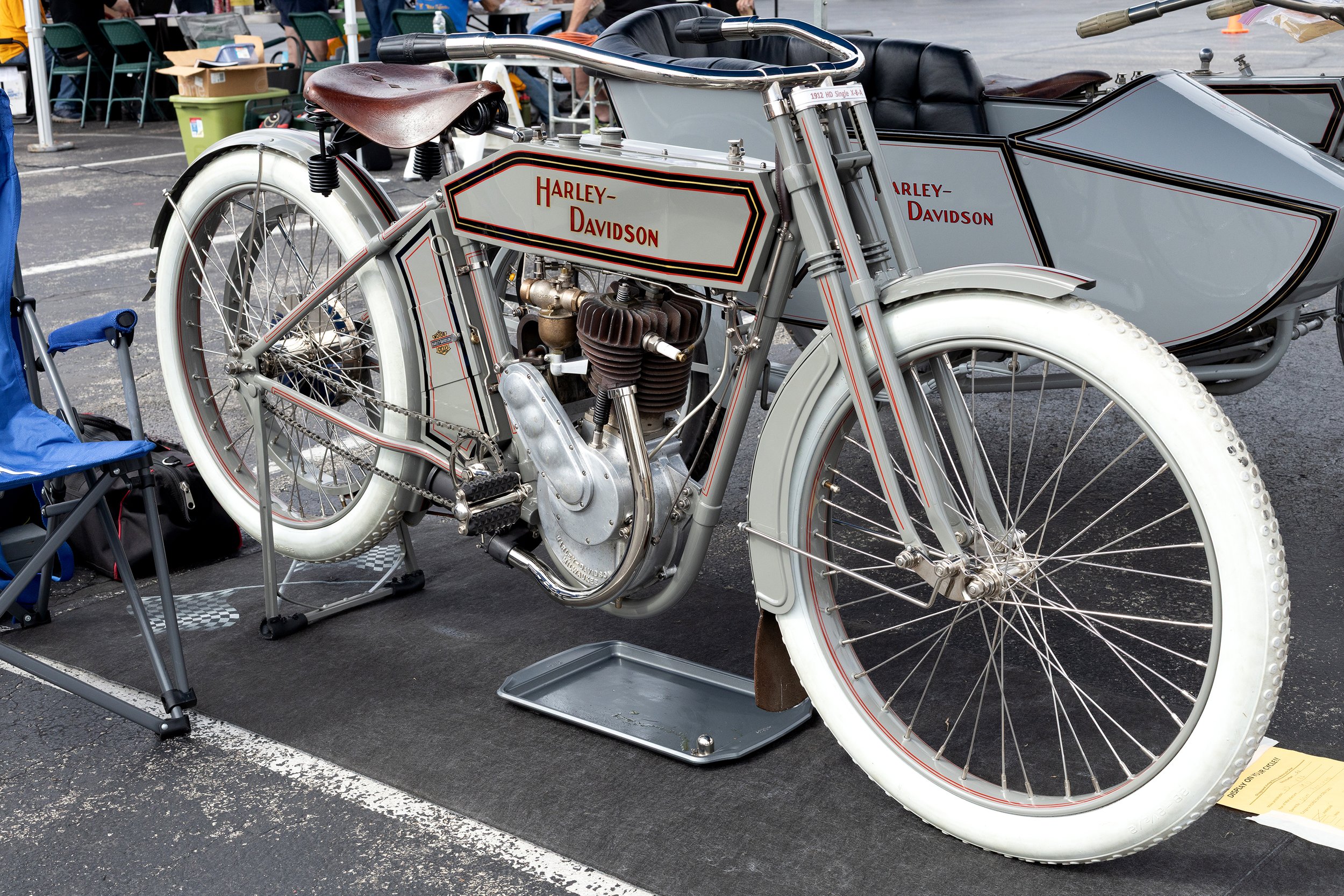 A vintage Harley-Davidson motorcycle with a sidecar on display at an outdoor event, surrounded by chairs, bags, and people.