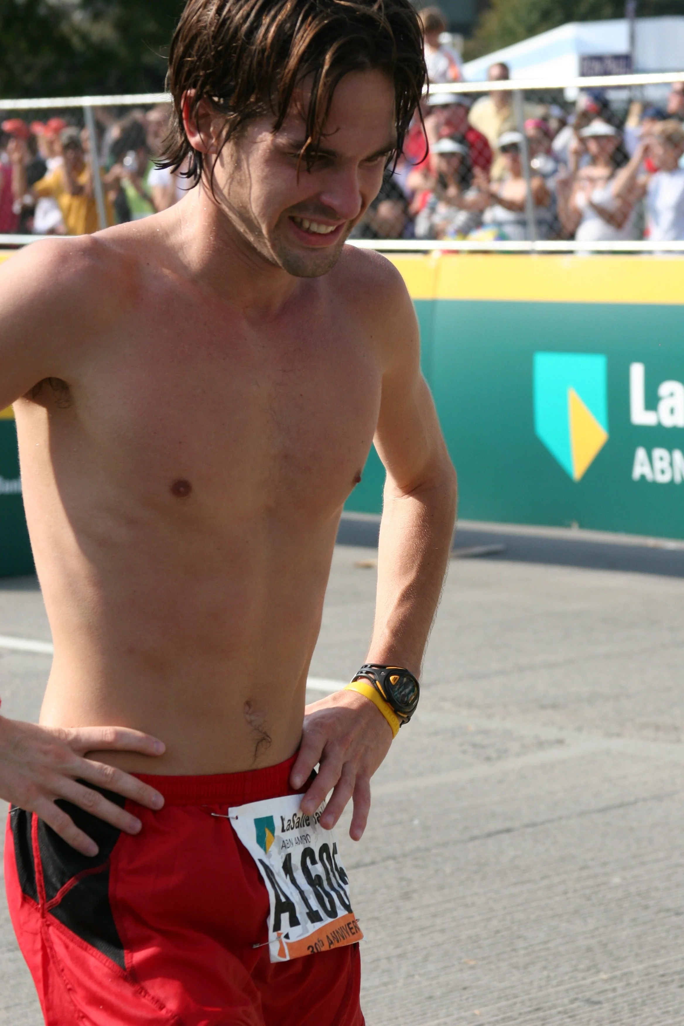 A male marathon runner with dark hair, shirtless, wearing red shorts and a wristwatch, standing after race, with a crowd of spectators in background.