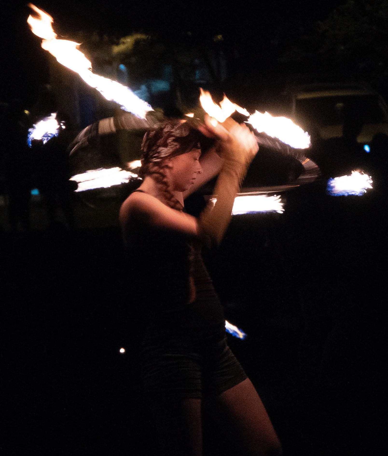 A person with long braided hair, dressed in black, performing a fire dance at night. They are holding a lit poi with flames, creating streaks of fire in the dark background.