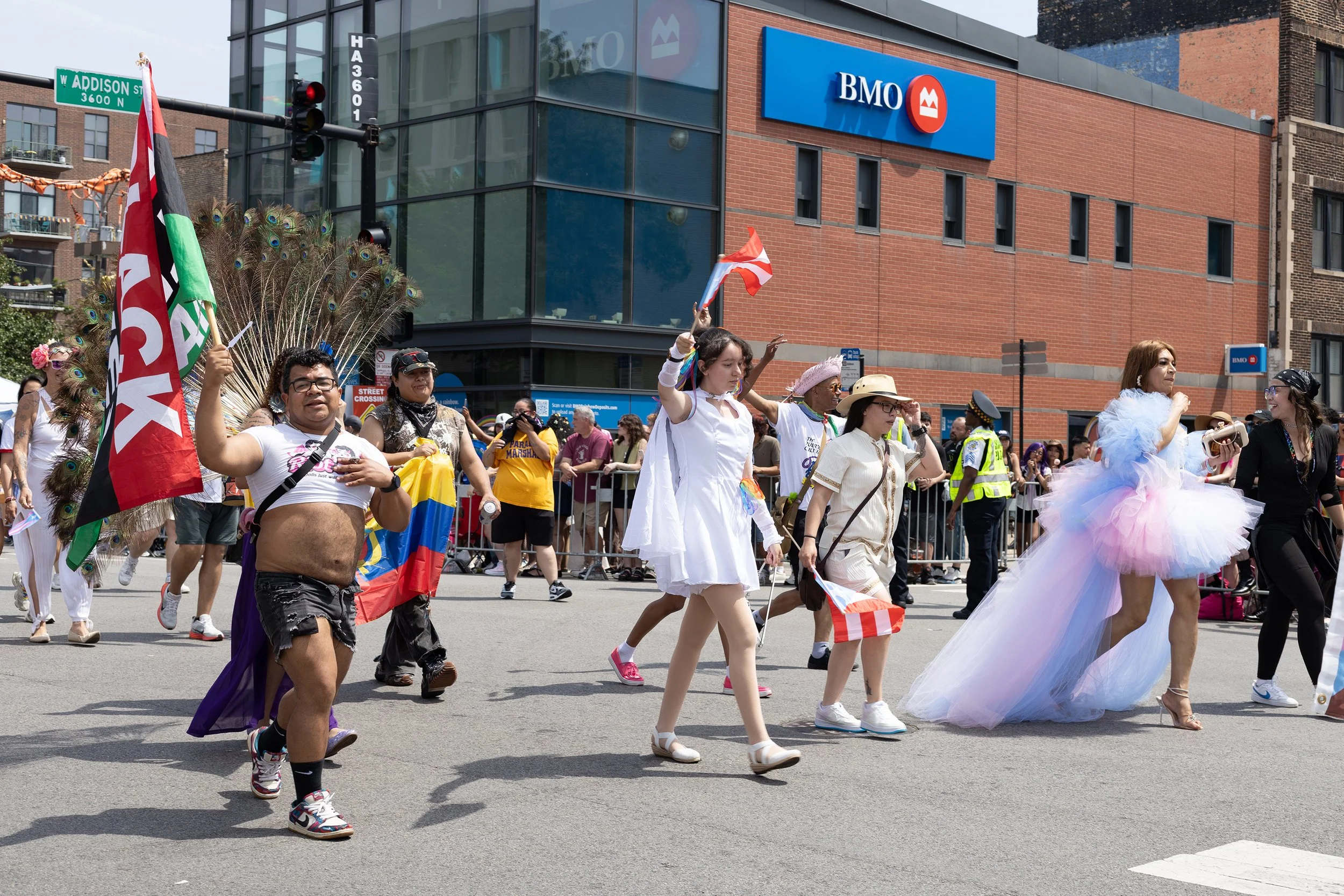 People participating in a parade or pride march on a city street, with some dressed in colorful costumes and holding flags, including LGBTQ pride flags. A large building with a BMO bank sign and a car traffic light are visible in the background.