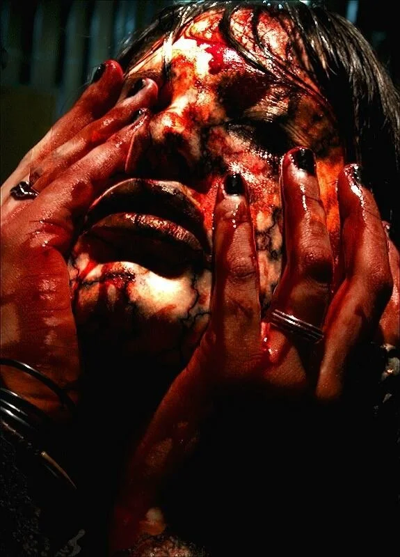 A person covered in blood, with their hands on their face and eyes closed, in a distressed state.