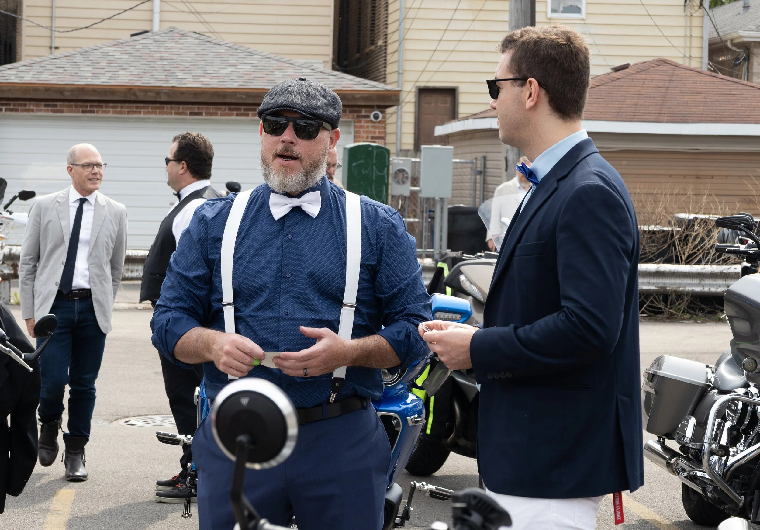Two men in suits and sunglasses engaged in conversation on a street with other men in the background, some also in suits, and parked motorcycles.