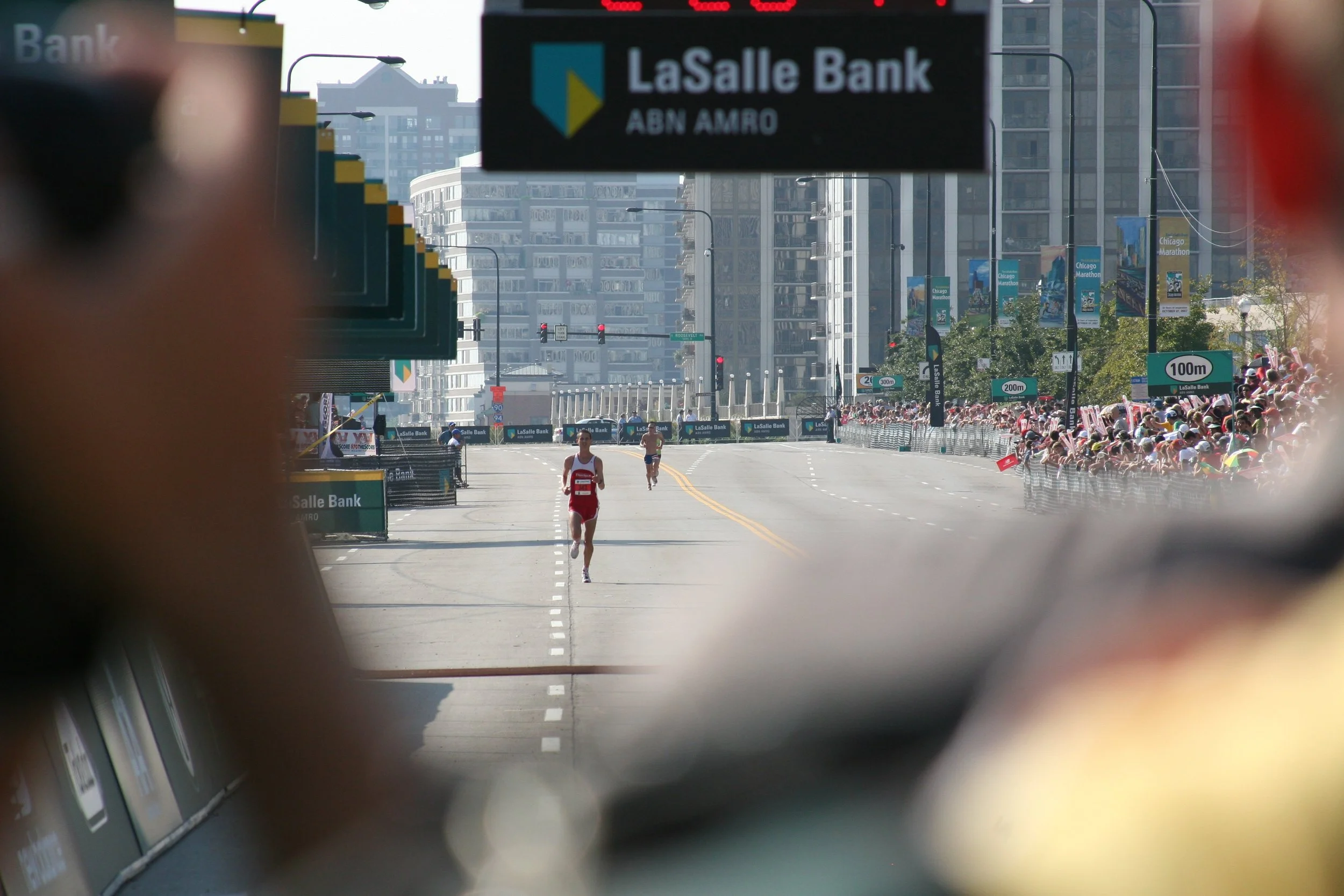 View of a city street during a marathon, with runners crossing the finish line, spectators on the side, and tall buildings in the background, as seen through a window or a cutout.