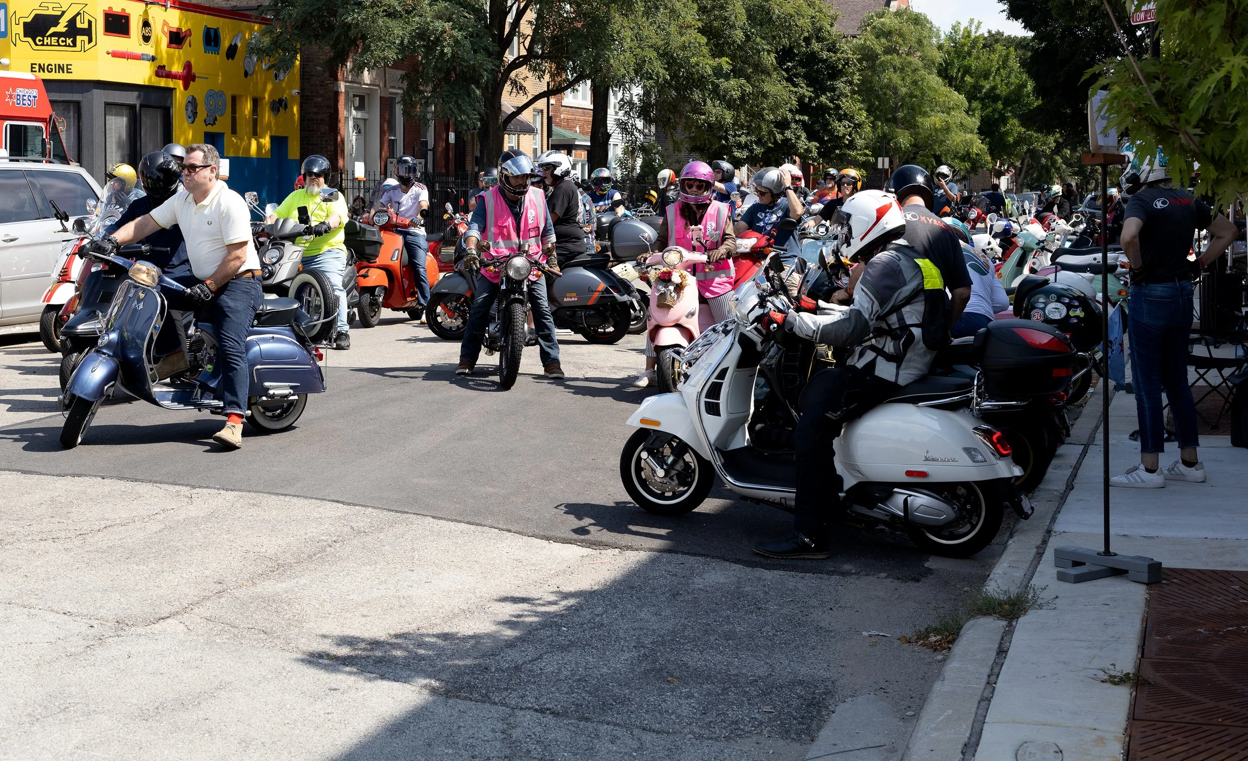 A crowd of people, many of whom are wearing helmets, gathered on a street with numerous motorcycles and scooters parked or moving. Some individuals are wearing pink vests, possibly indicating they are organizers or guides. There is a colorful buildin