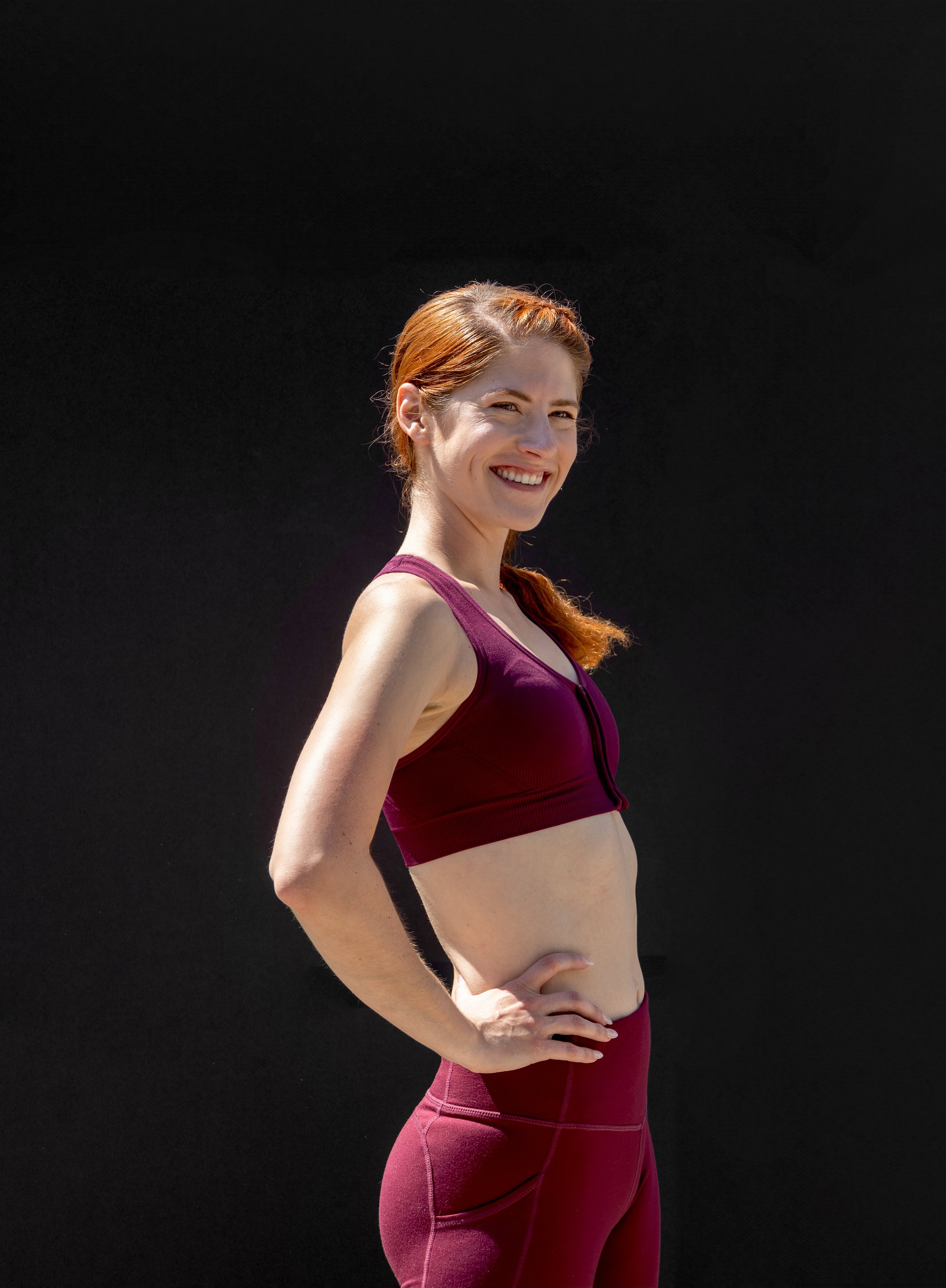 A woman with red hair in workout clothes, smiling with her hand on her hip, standing against a black background.