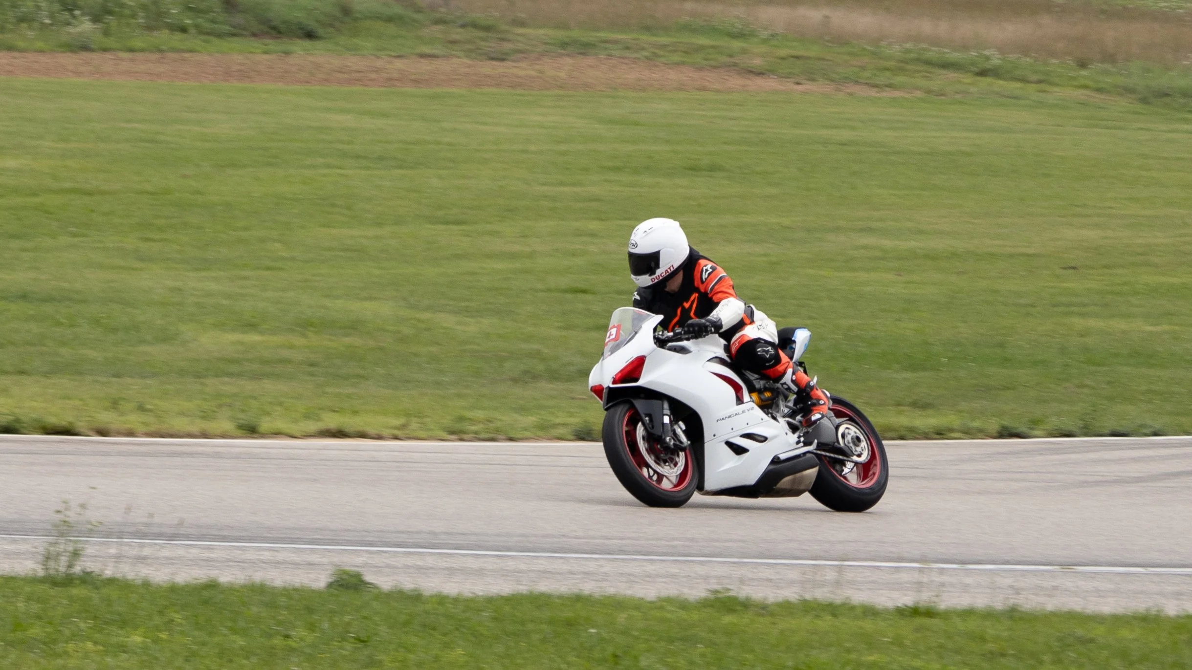 A motorcyclist in black and orange riding gear and a white helmet riding a white and red sport motorcycle on a paved road, with green grass and a slight hill in the background.