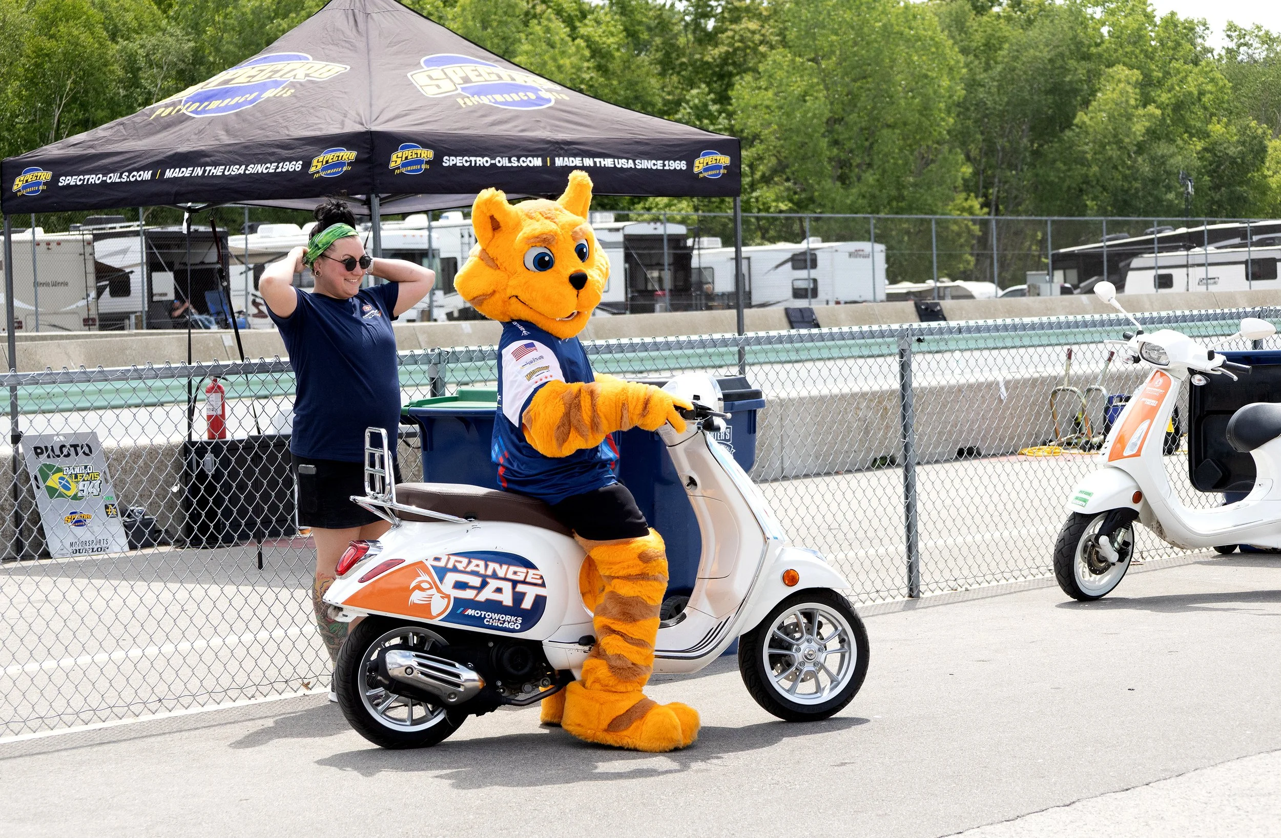 A person in a blue shirt and green headband stands beside a white scooter with an orange cat mascot riding it. The mascot has large blue eyes, a big smile, and is dressed in a blue sports jersey. They are near a chain link fence with a racetrack and 