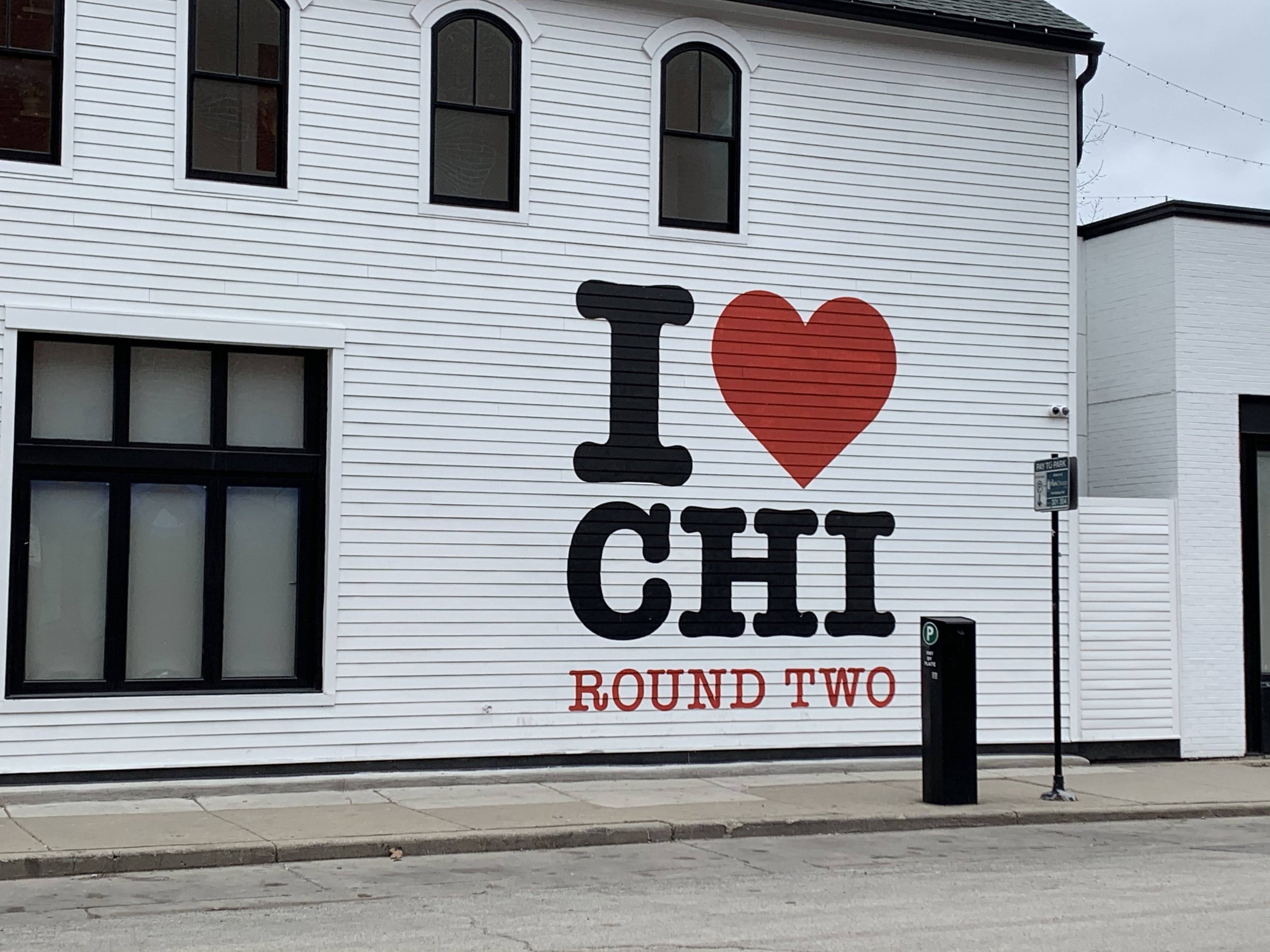 A white building with three black-framed windows and a large sign that reads "I ♥ CHI" with the text "ROUND TWO" in red at the bottom. There is a parking sign and pole in front of the building, and a sidewalk and street at the foreground.
