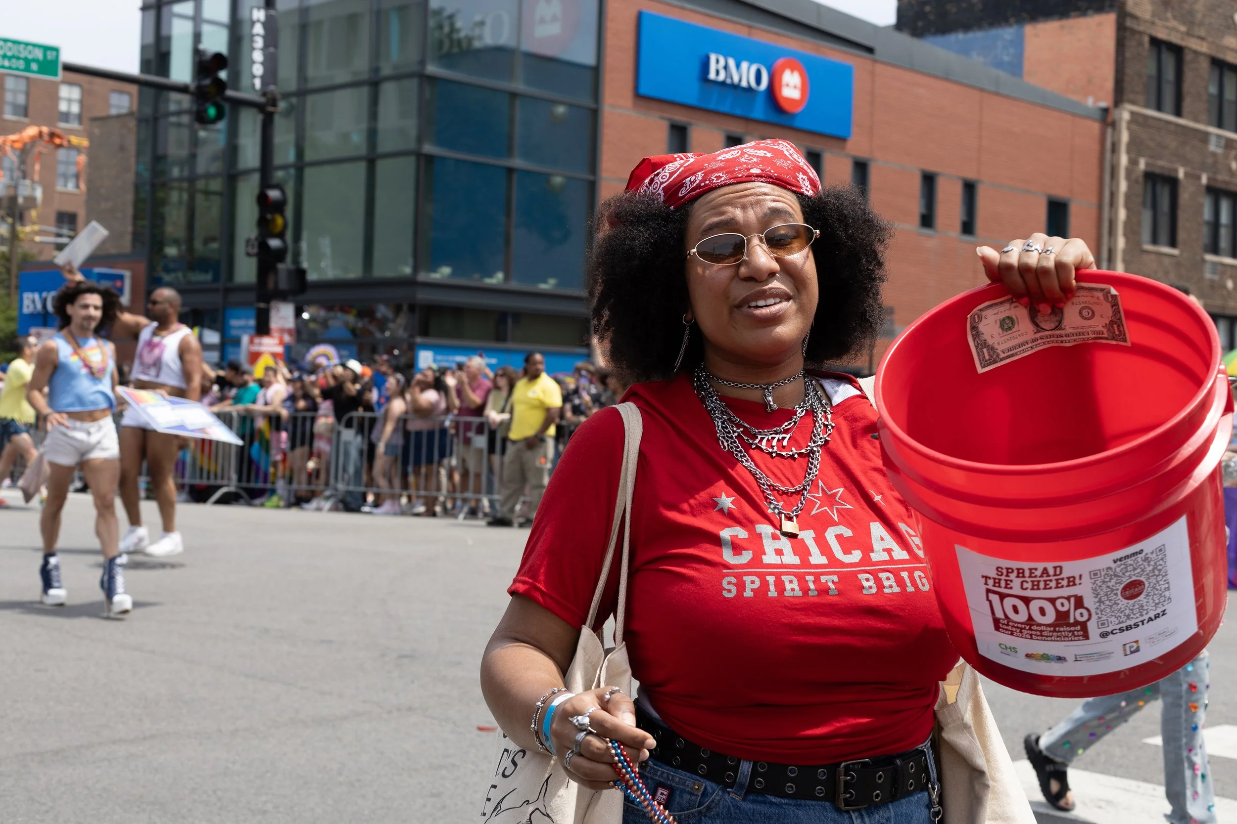 A woman holding a red bucket with a dollar bill inside at a Pride parade, with a crowd, rainbow flags, and a BMO bank building in the background.