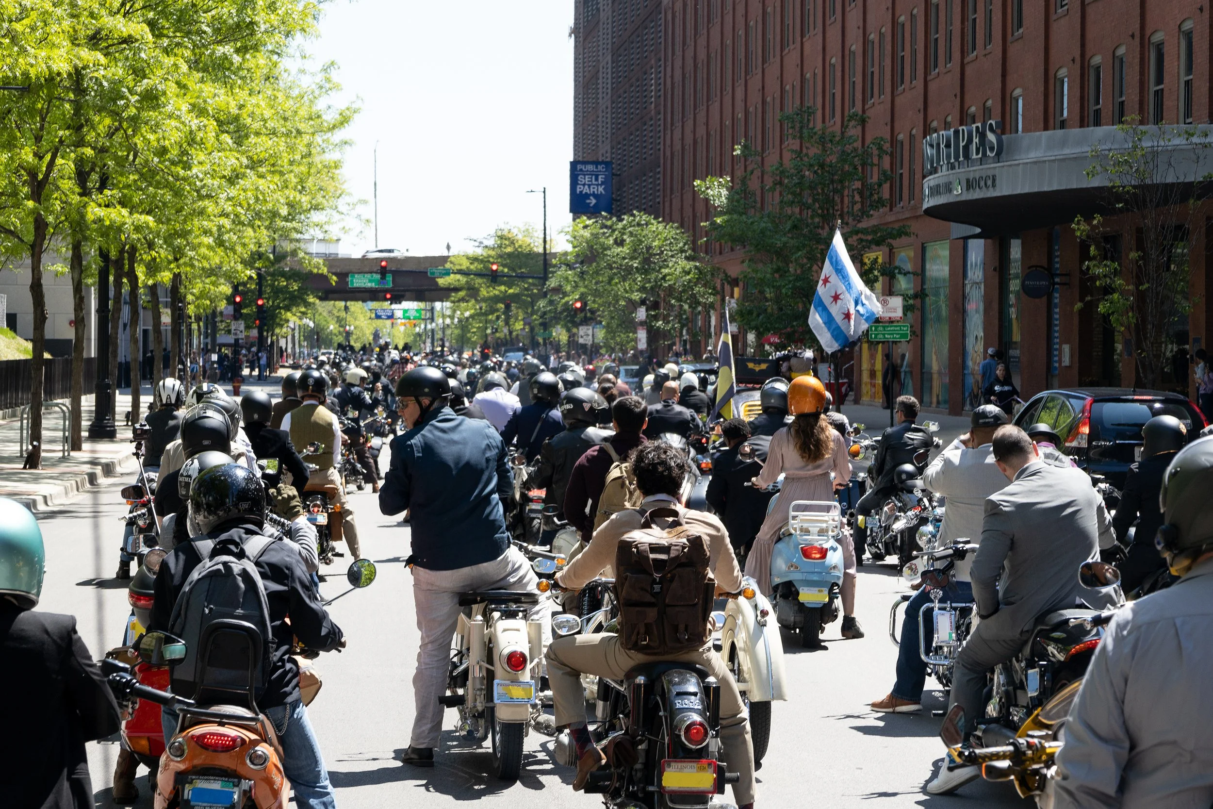 A busy city street filled with people riding and standing around on motor scooters. Green trees line the sidewalk, and tall red brick buildings are visible on the right. Traffic lights and signs are overhead, and a Chicago flag is being waved by one 