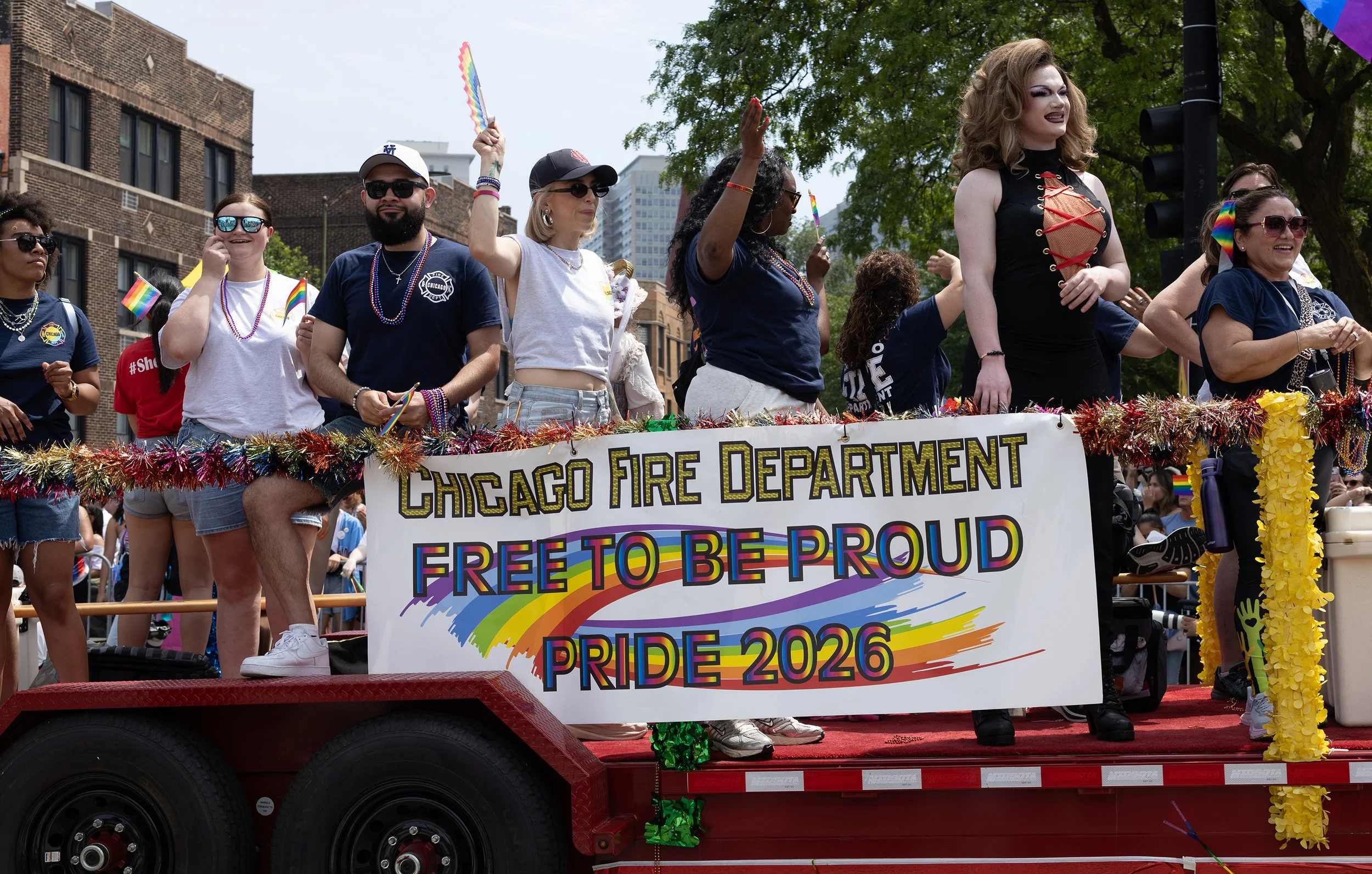 People standing on a float in a parade, holding rainbow flags and wearing festive attire. The float has a sign that reads 'Chicago Fire Department Pride 2026' with a rainbow design.