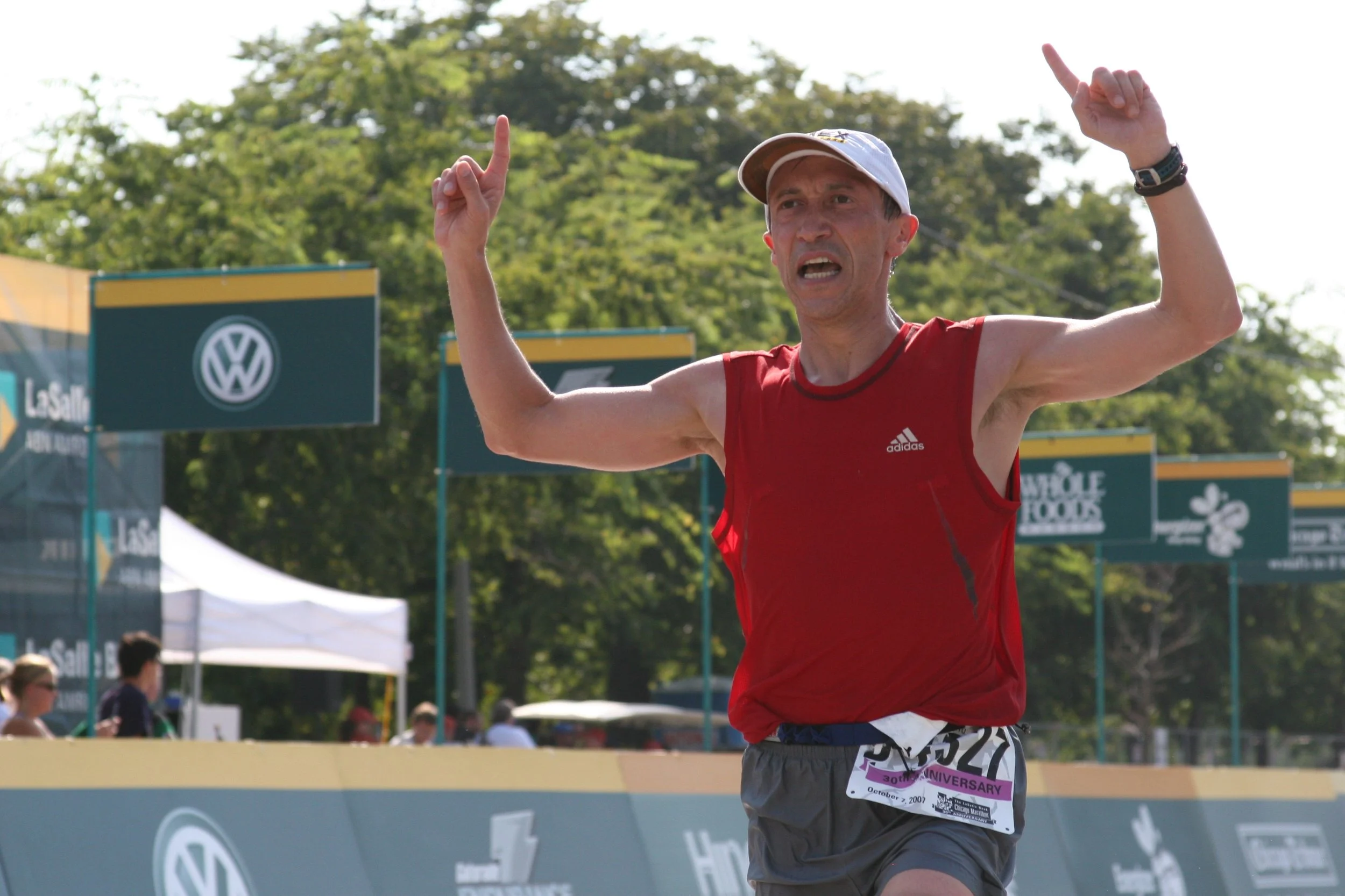 Male runner in a red tank top and gray shorts, wearing a white cap, crossing the finish line of a race outdoors, with trees and sponsor banners in the background.