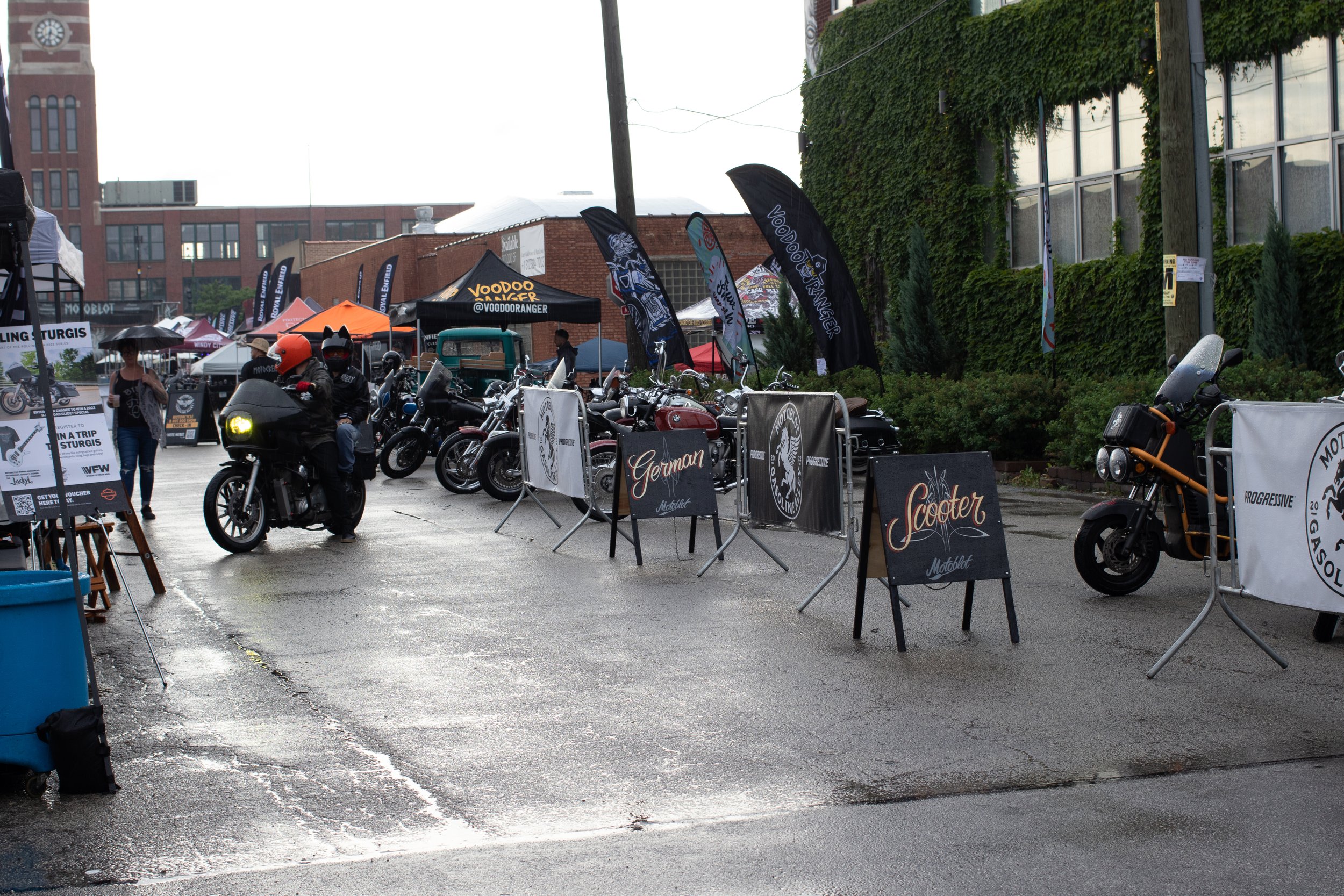 Outdoor motorcycle and scooter exhibition with banners, tents, and people, including some wearing helmets, on a rainy day.