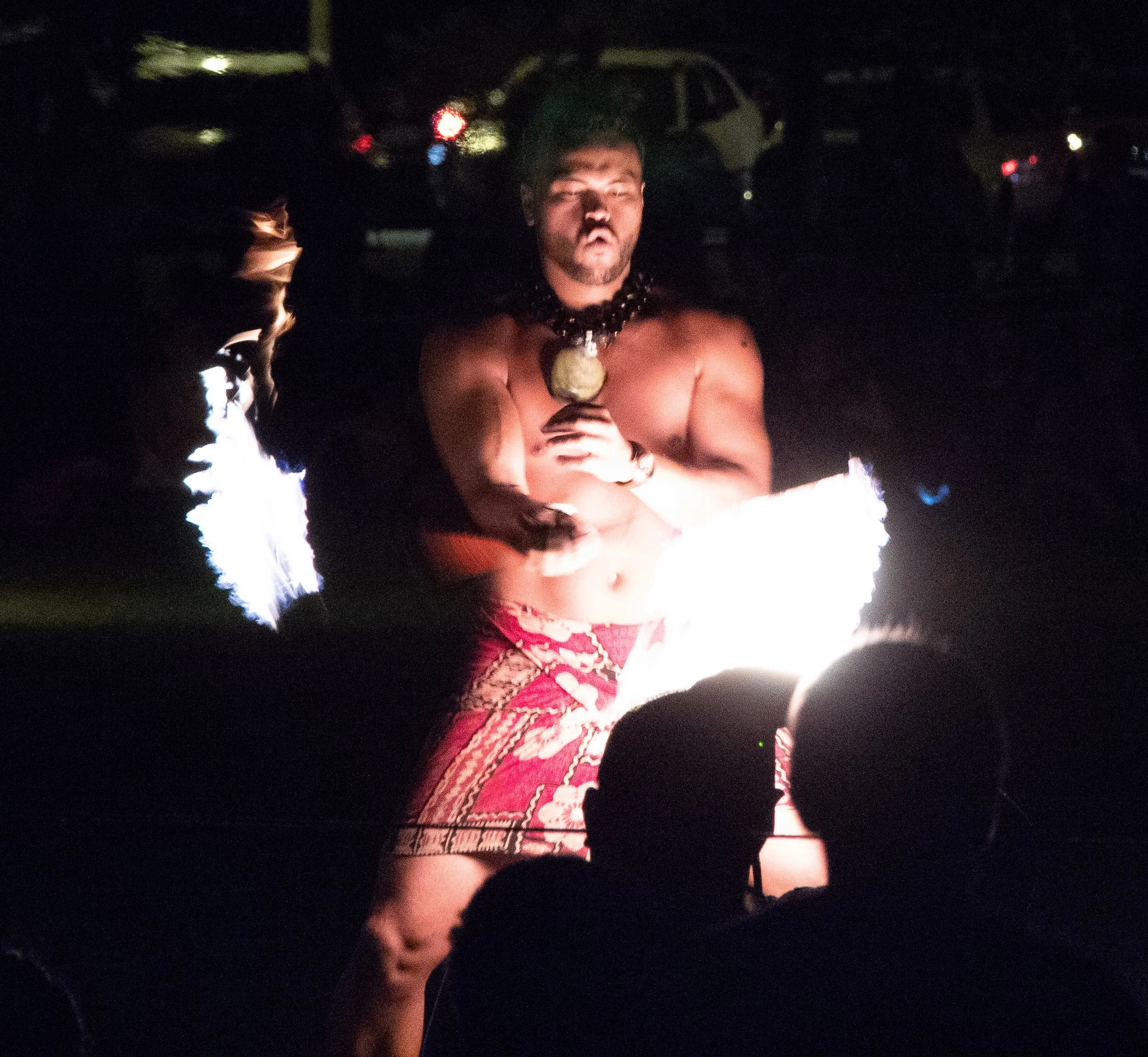 A shirtless man with a necklace holding a flaming object at night in a dark outdoor setting. His expression is serious and he appears to be participating in a fire performance.