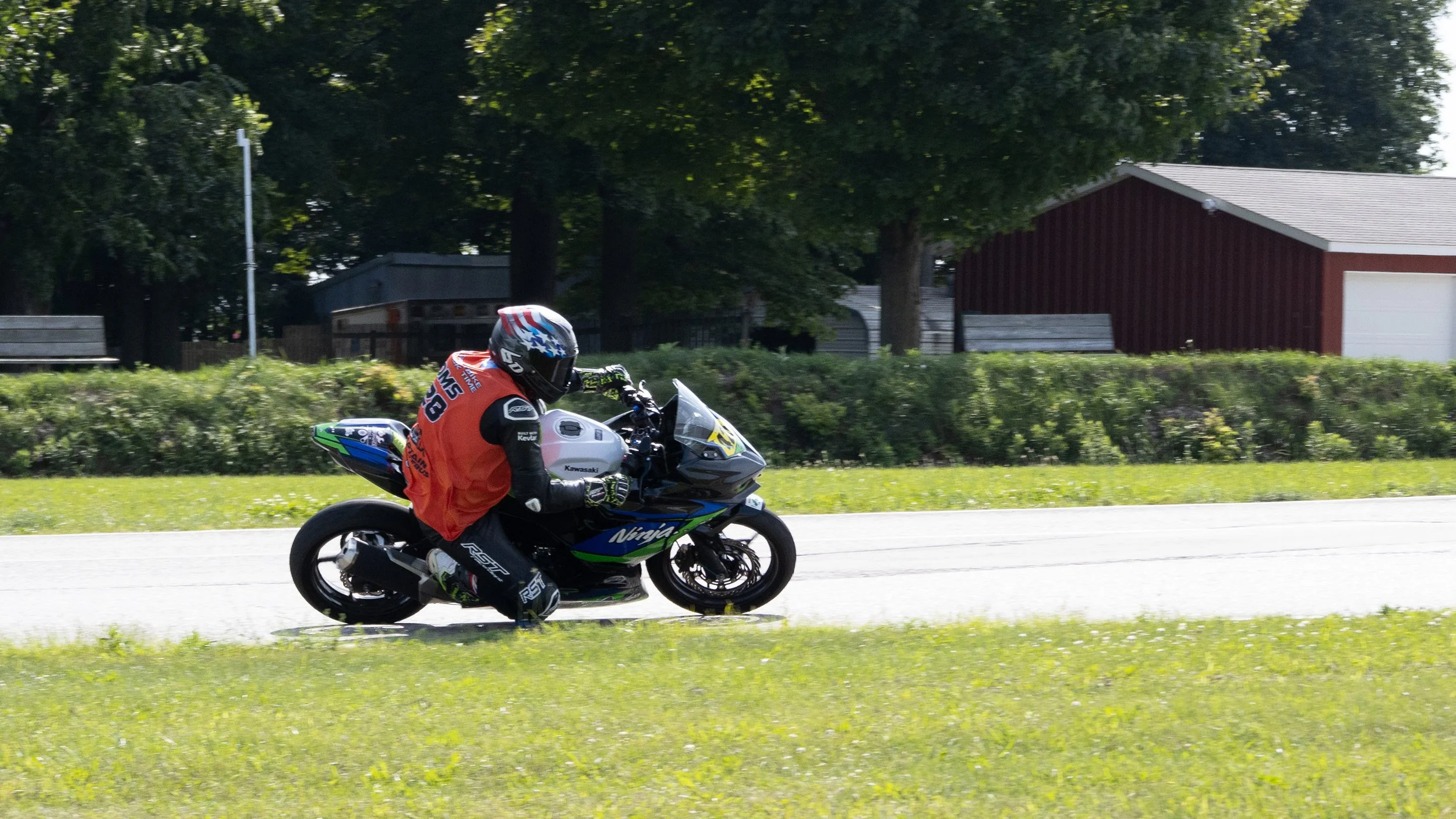 A motorcyclist riding a black and green Kawasaki Ninja motorcycle on a paved road in a rural area with green grass, trees, and houses in the background. The rider is wearing a helmet, gloves, and a red vest.