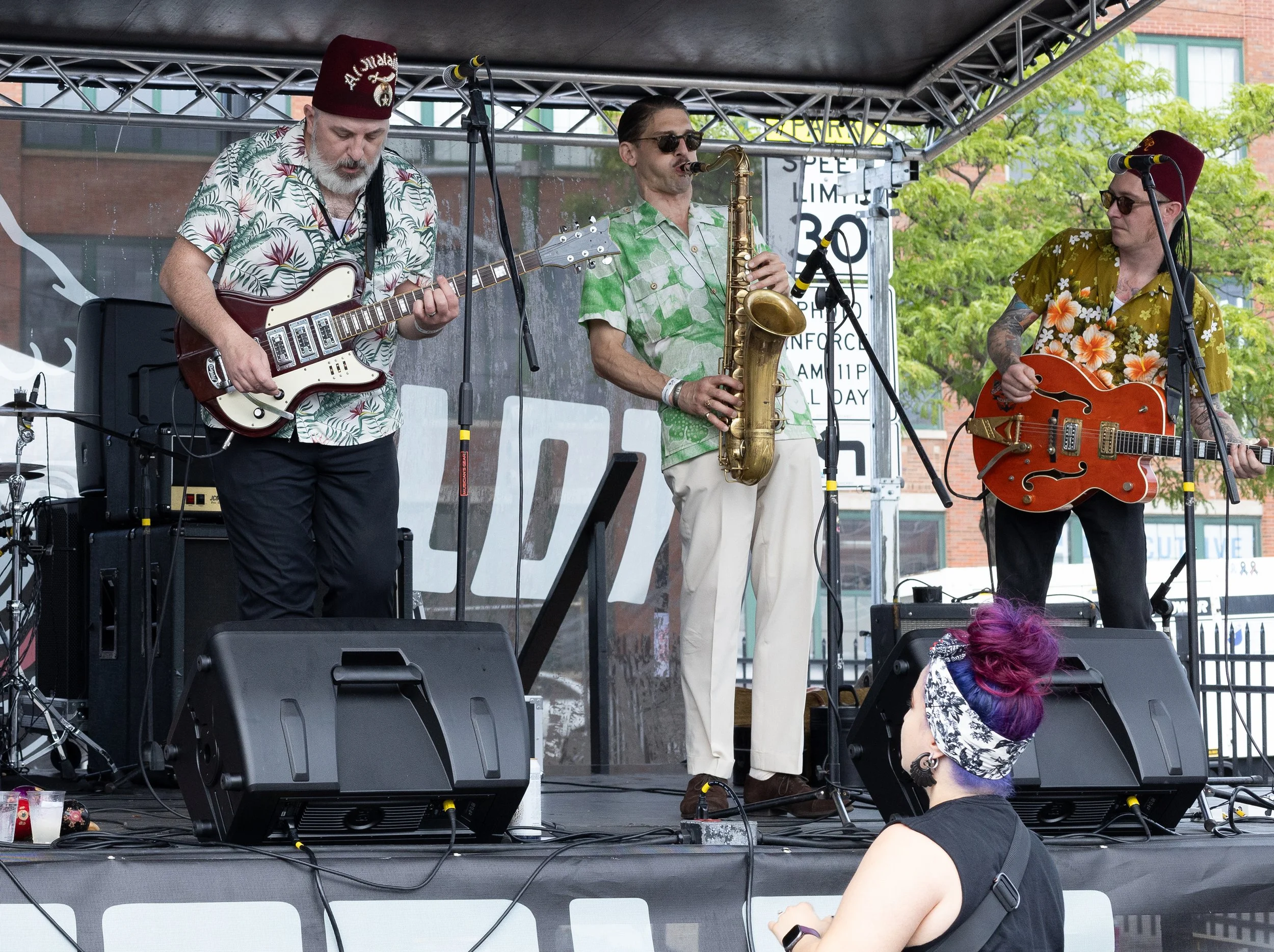 Three band members performing on an outdoor stage. The guitarist on the left wears a floral shirt and a red fez hat. The saxophonist in the center wears a green shirt and sunglasses. The guitarist on the right wears a yellow floral shirt and a red fe