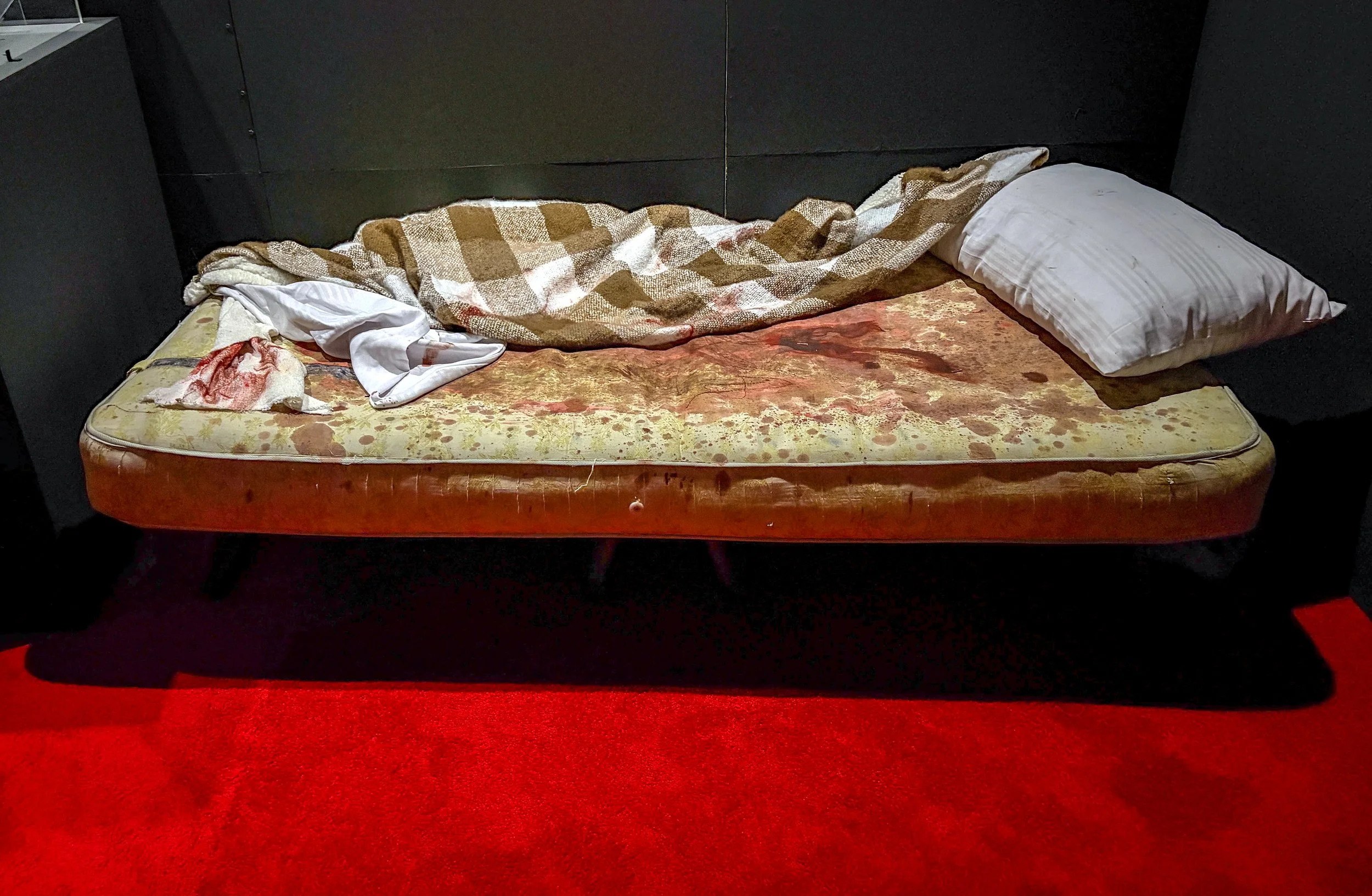 A mattress on black bed frame with stained sheet, white pillow, blanket, and some cloth lying on it, with red carpet underneath.