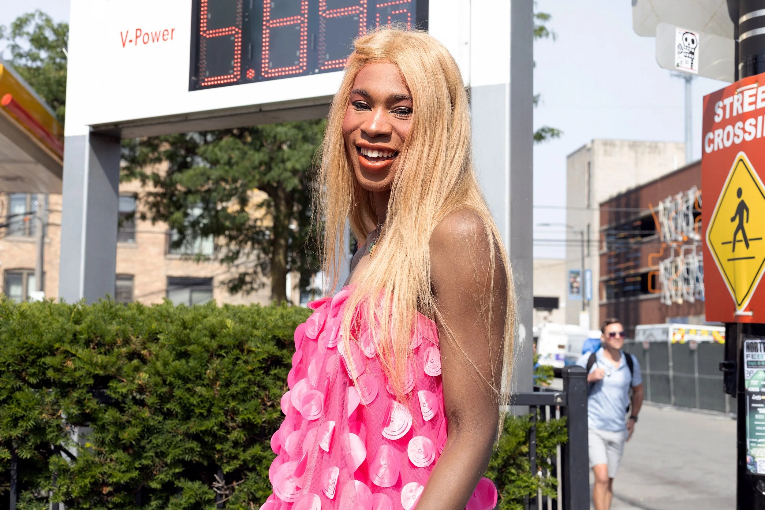 A smiling woman with long blonde hair wearing a pink dress with large fabric flowers walks outdoors on a city street with traffic signs, a digital speed display, and a few pedestrians in the background.