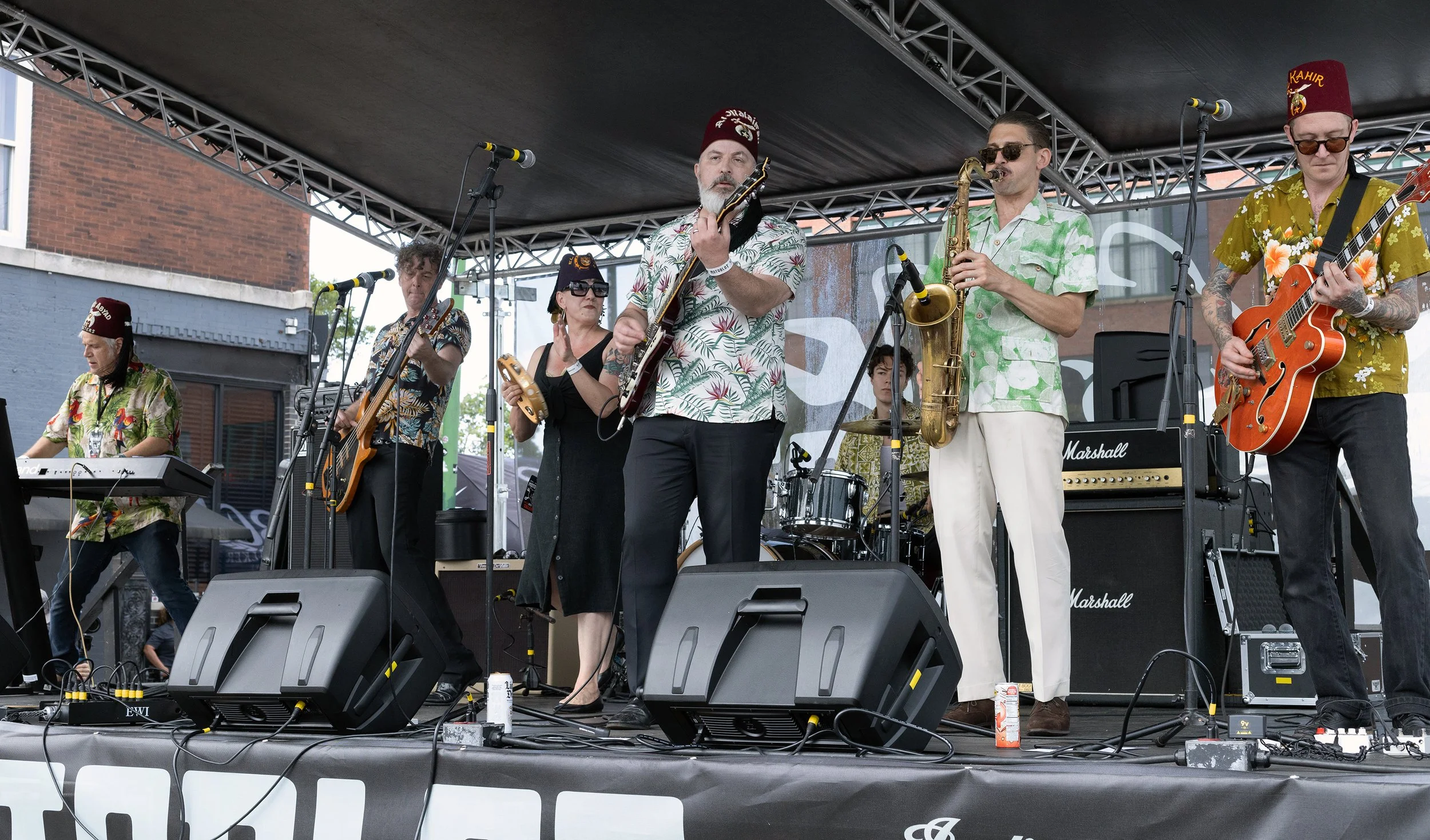 A band performing live on outdoor stage, featuring musicians with guitars, saxophone, keyboard, and a drummer, all dressed in tropical-themed shirts and some wearing red fez hats.