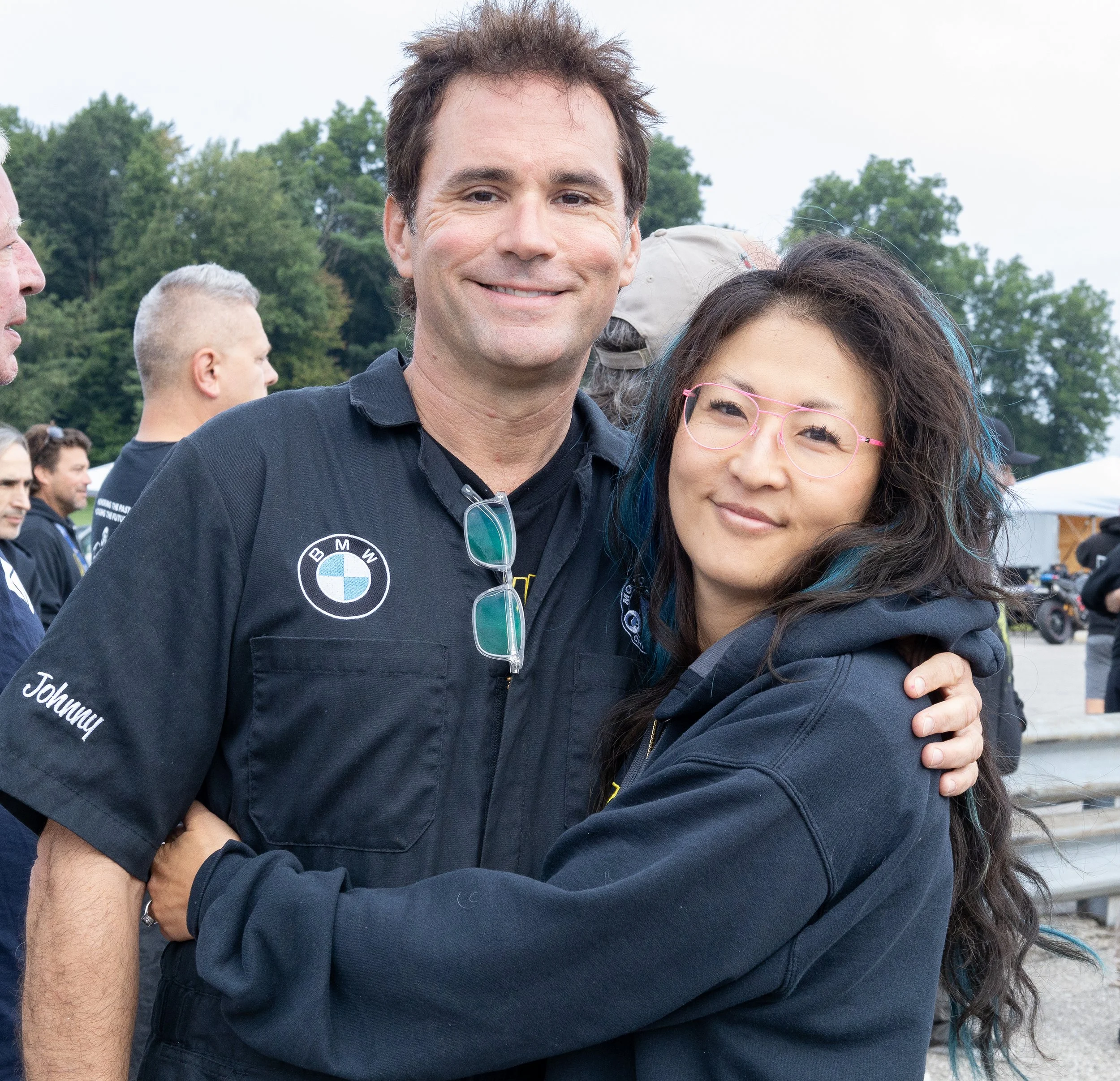 Two people hugging outdoors with trees and a cloudy sky in the background.
