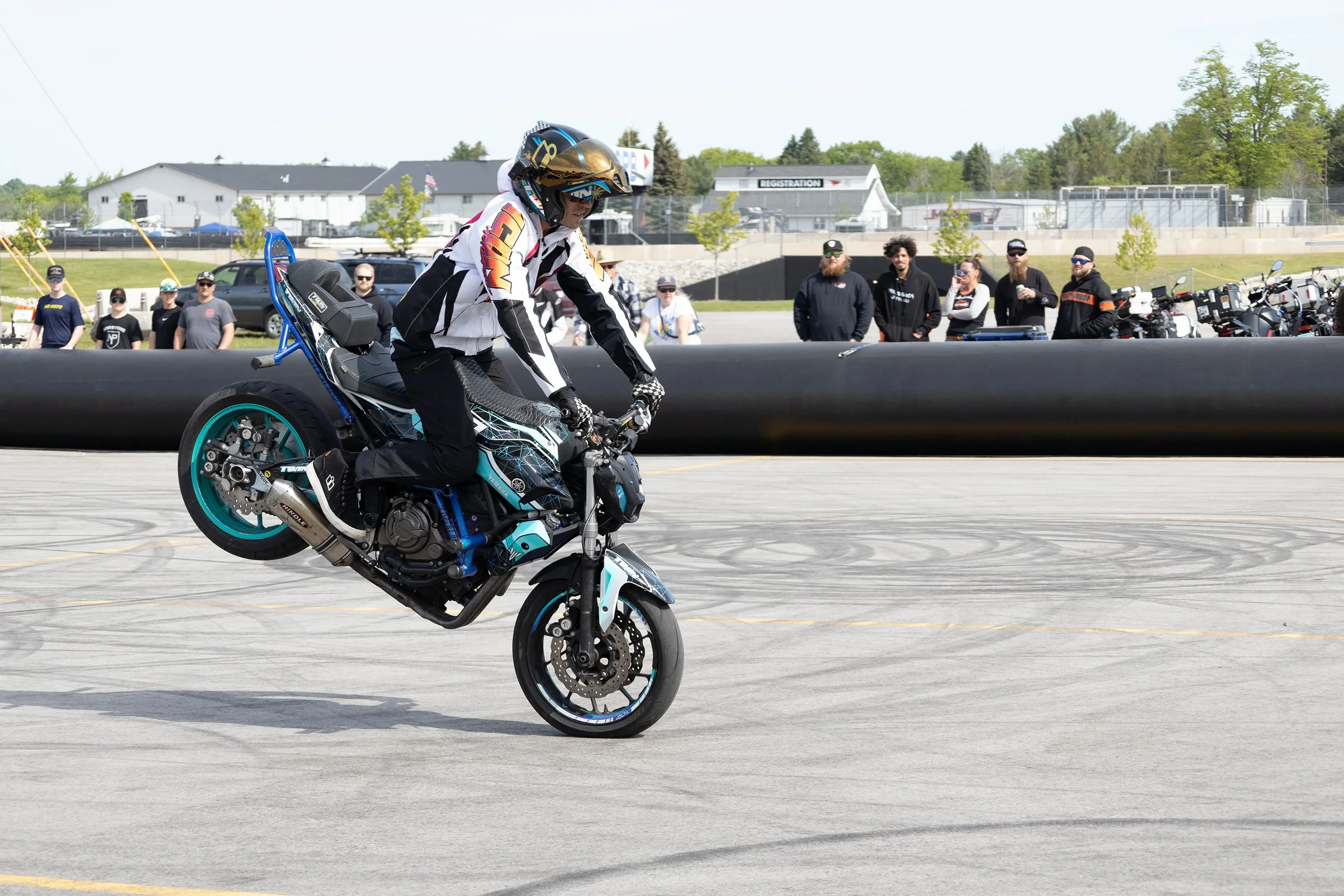 A person in a black and white racing suit riding a Yamaha motorcycle on a paved area with a group of people and motorcycles in the background.