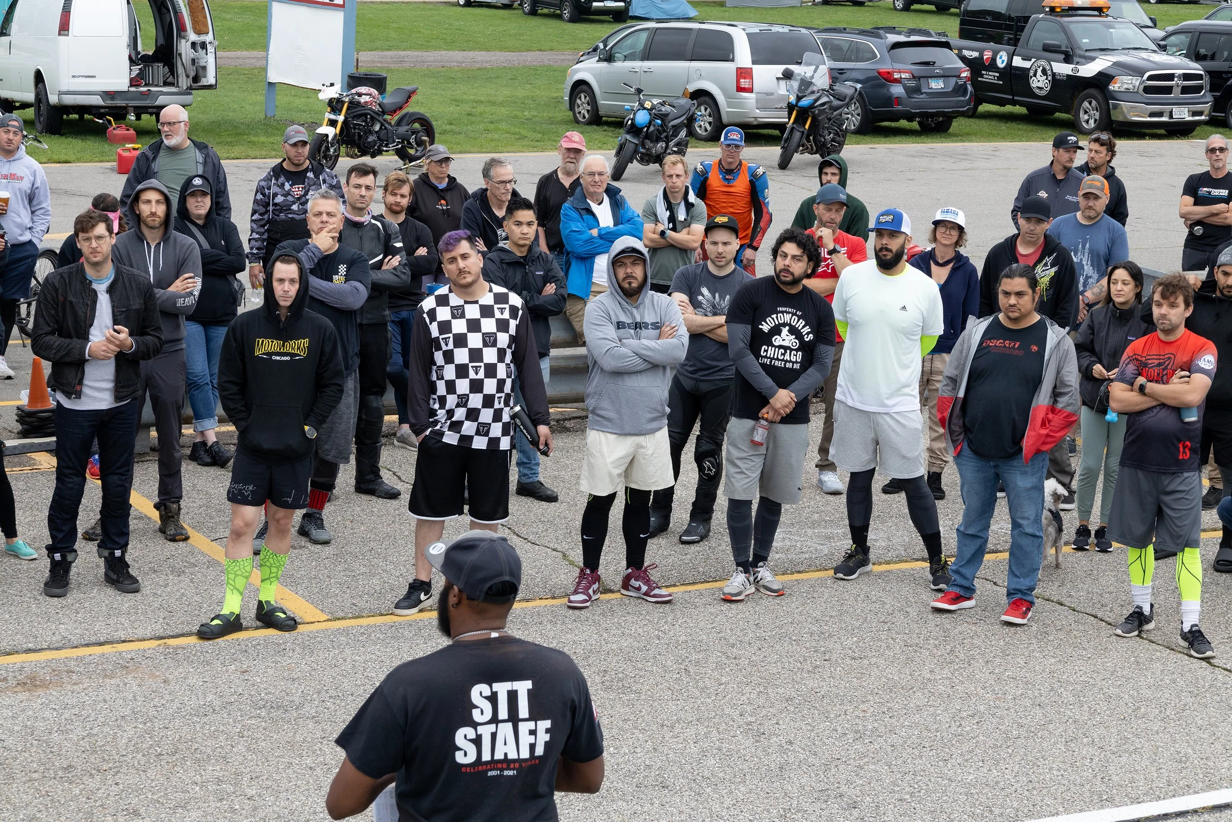 Group of people gathered outdoors, some dressed in motorcycle gear, standing on a parking lot with parked cars and motorcycles in the background.